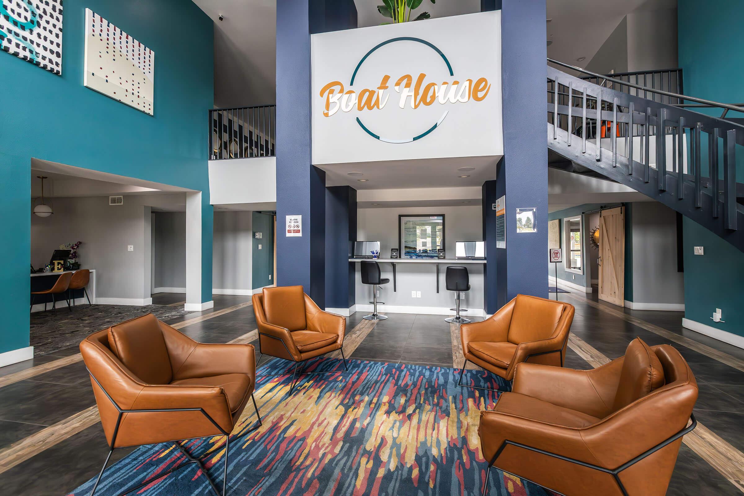 A modern lobby area featuring four stylish brown leather lounge chairs arranged around a colorful area rug. The walls are painted in a deep teal color, and there's a large sign that reads "Boat House" on the wall. A reception desk is visible in the background, with seating and decor elements enhancing the contemporary aesthetic.