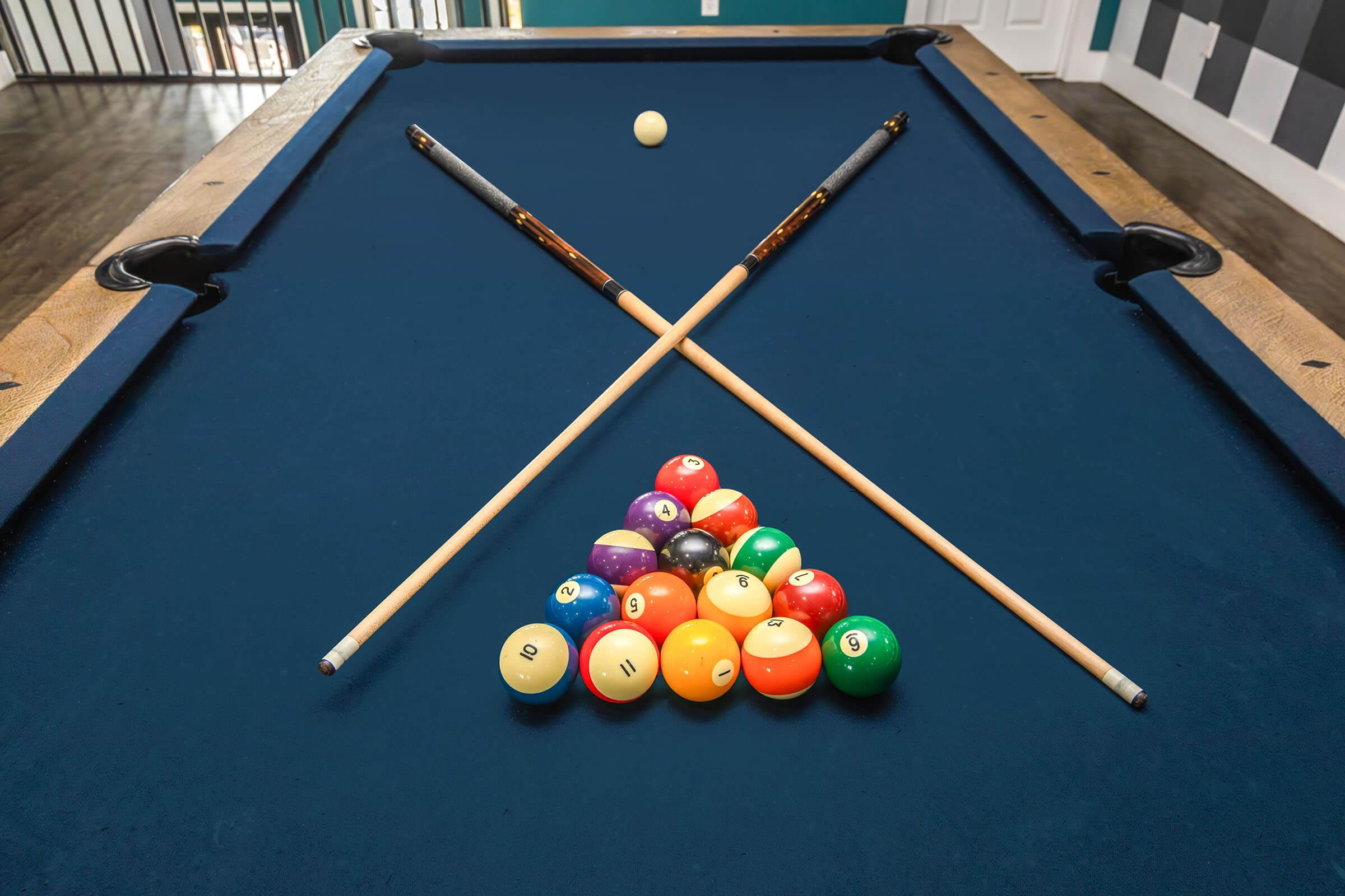 A pool table with a triangle of colorful billiard balls arranged at the base. Two cue sticks are crossed above the triangle, and a white cue ball is positioned nearby. The table has a blue felt surface, and there are wooden edges. The background shows a room with a door and patterned wall.