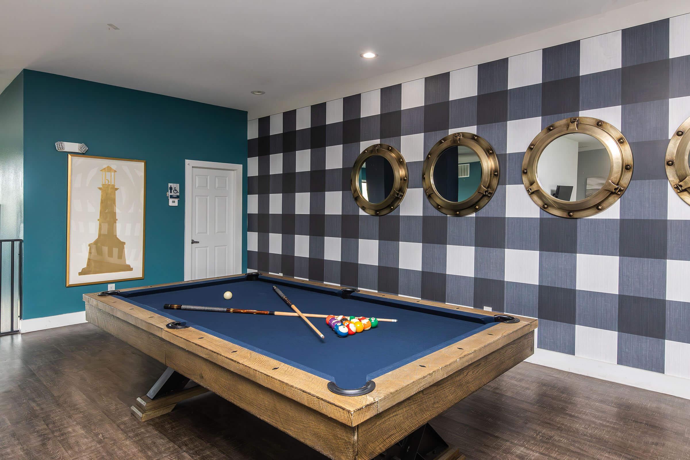 A modern game room featuring a billiards table with a blue felt surface, cue sticks, and colorful balls. The walls are decorated with alternating blue and grey checkered patterns, and there are circular mirrors hanging on the wall. A framed picture of a lighthouse is also visible.