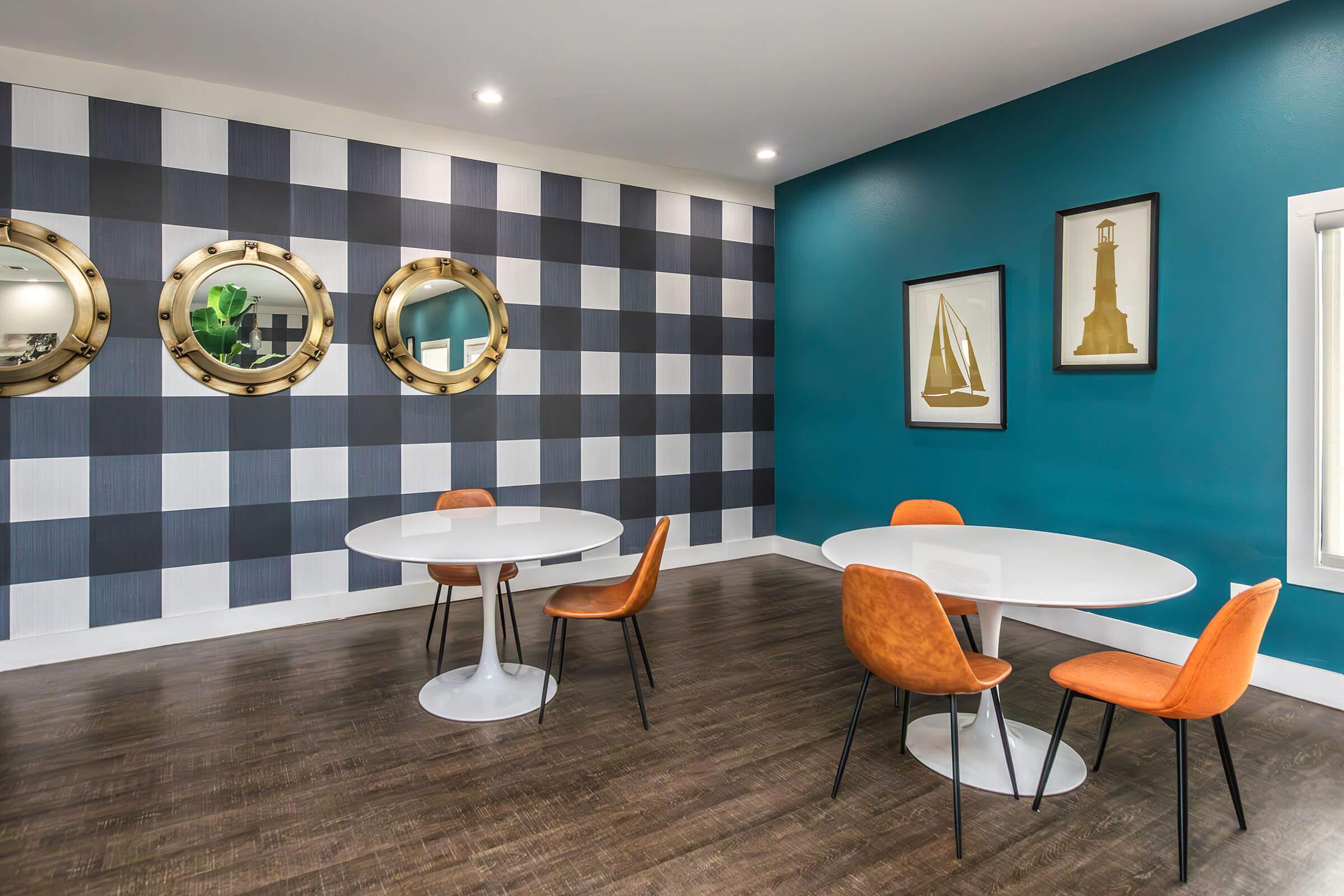 Modern room featuring two round white tables with orange chairs, set against a stylish checkered wall and a contrasting teal wall. The decor includes gold-framed mirrors and framed artwork depicting a sailboat and a lighthouse. The space has a clean, contemporary look with warm wood flooring.