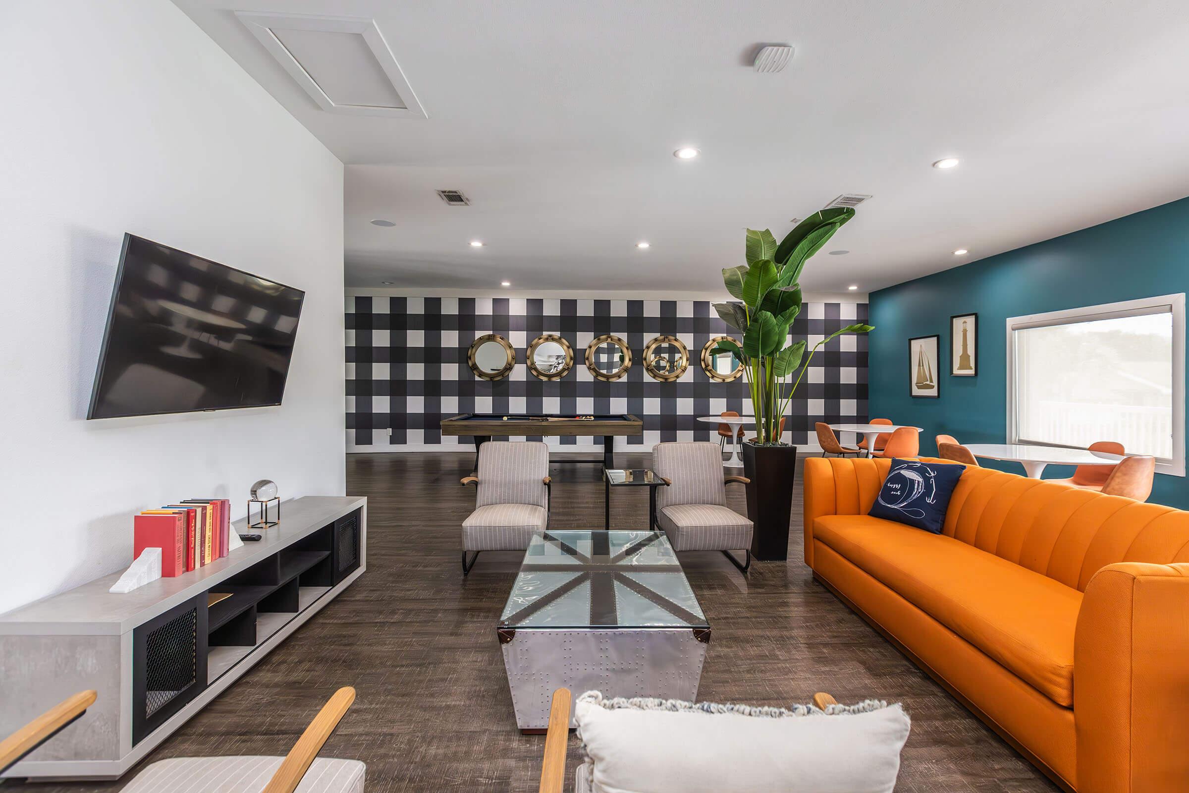 A modern, stylish lounge area featuring an orange couch, contemporary chairs, and a coffee table. There is a large TV mounted on the wall, and a striking checkered wallpaper. Decorative mirrors adorn the wall, with indoor plants adding a touch of greenery to the space. Soft lighting enhances the inviting atmosphere.