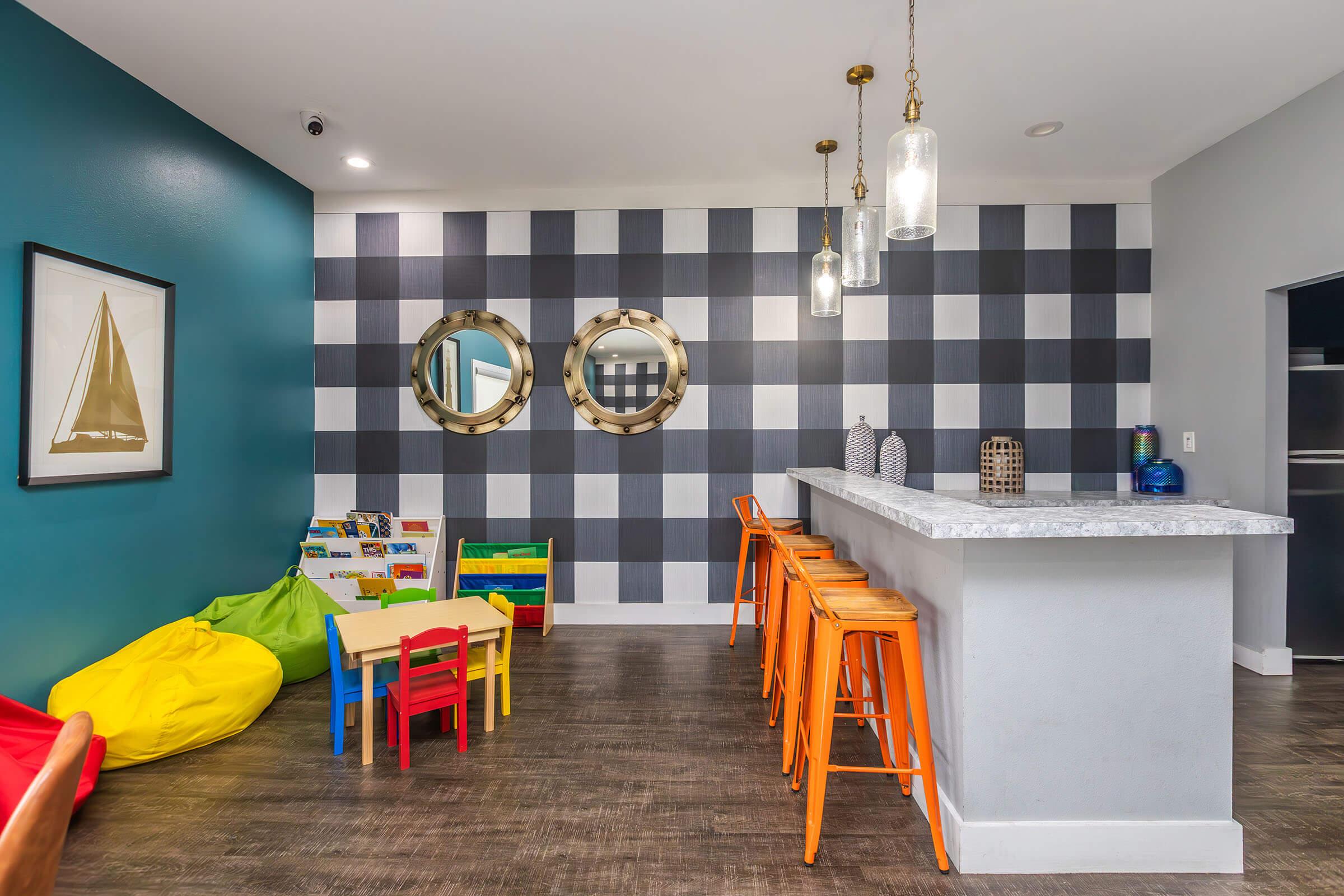 A bright and modern playroom featuring colorful bean bags, a small table with chairs, and a bar area with orange stools. The wall is decorated with large circular mirrors and a checkered pattern, accented by a framed nautical-themed artwork. The space is designed for children to play and engage creatively.