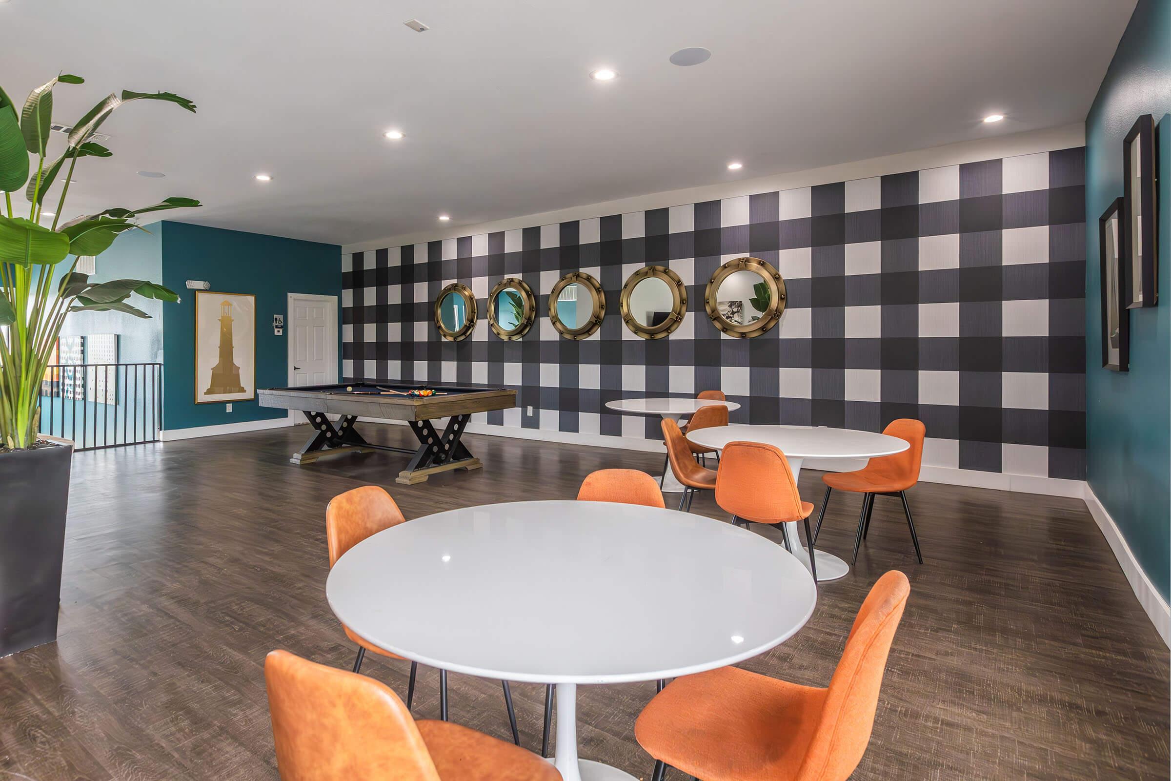 A modern recreational lounge featuring a pool table, round white tables with orange chairs, and a wall adorned with large mirrors. The decor includes a combination of blue and checkered patterns, along with indoor plants for a fresh ambiance. Natural light enhances the inviting atmosphere.