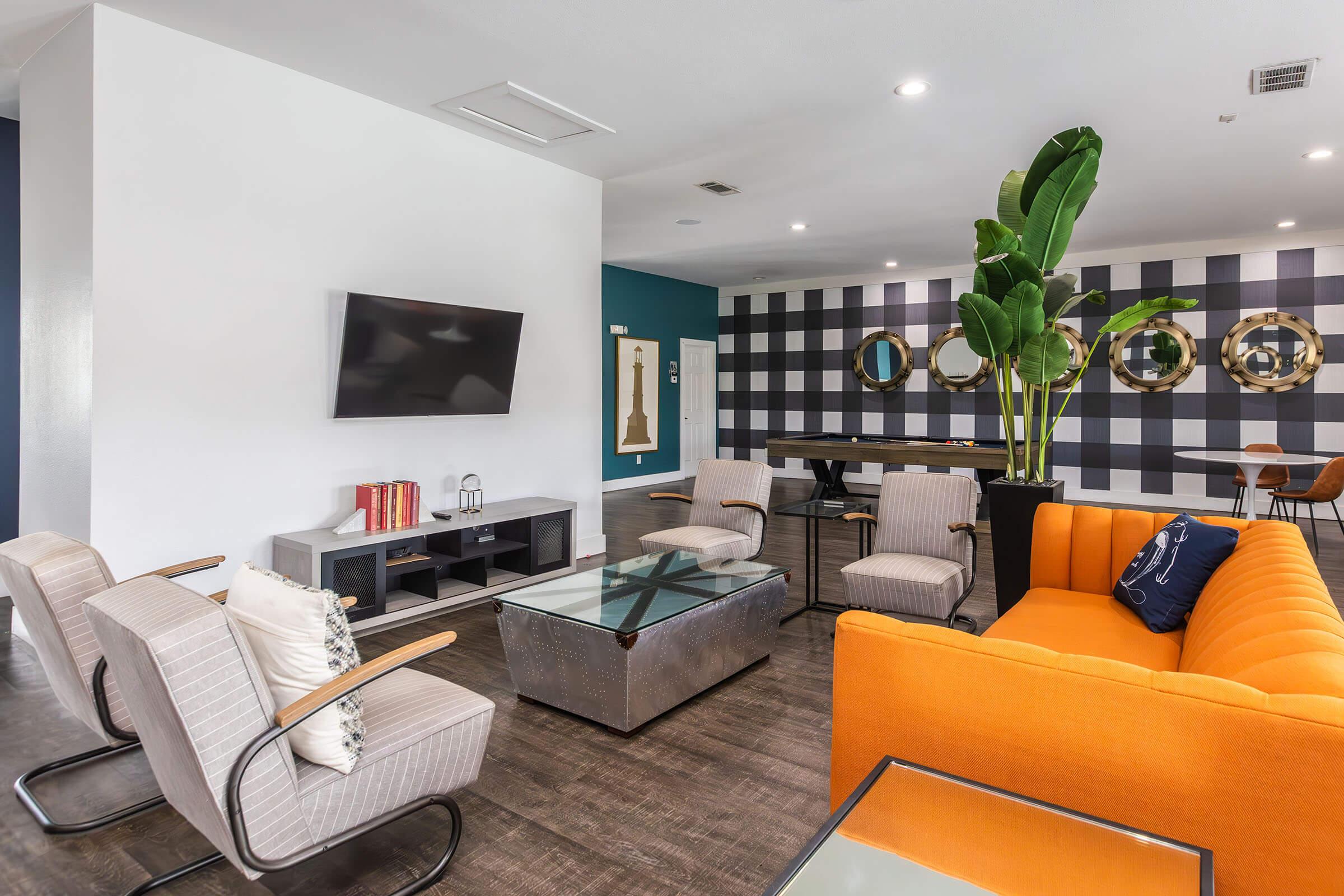 A modern living area featuring an orange sofa, a glass coffee table, and stylish chairs. The walls are decorated with a black and white plaid pattern and round mirrors. A large flat-screen TV is mounted on one wall, and a tall green plant adds a touch of nature to the space.