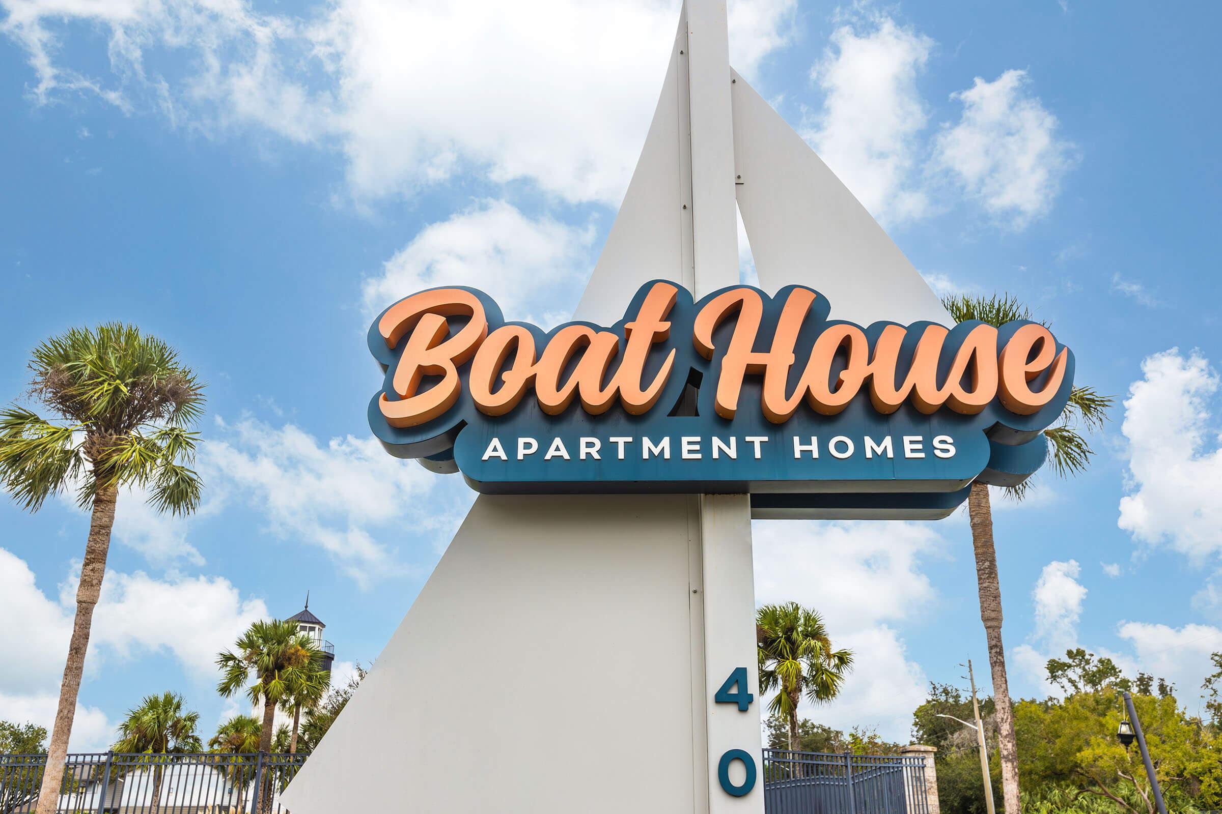 Sign for "Boat House Apartment Homes" featuring a stylized font, with palm trees in the foreground and a clear blue sky in the background. The sign is prominently displayed on a white structure designed to resemble a sail.