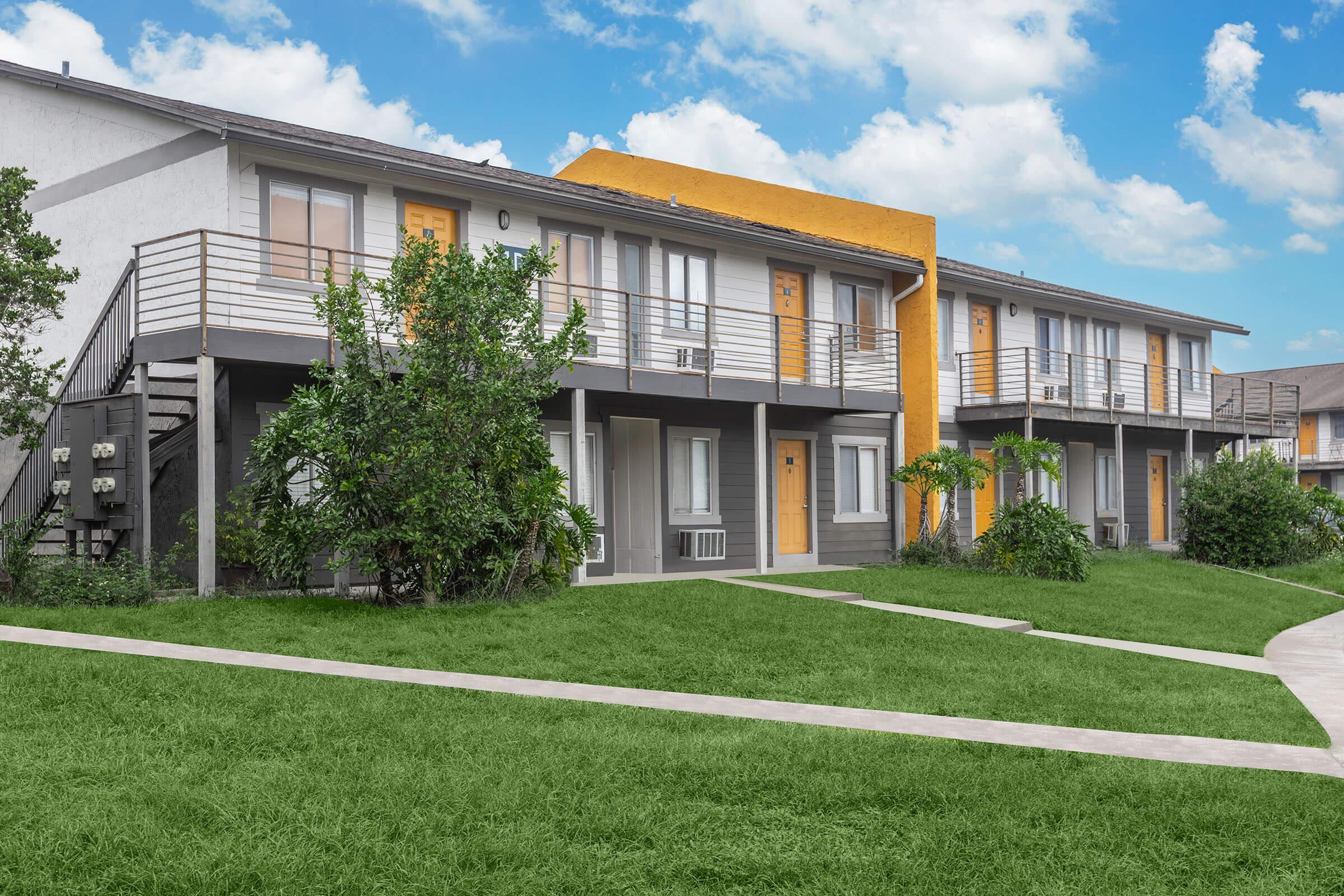 A modern apartment community featuring two-story buildings with a mix of gray and orange facades. Lush green lawns surround the buildings, and paths lead to individual entrances. The sky above is bright with fluffy clouds, adding to the inviting atmosphere of the residential area.