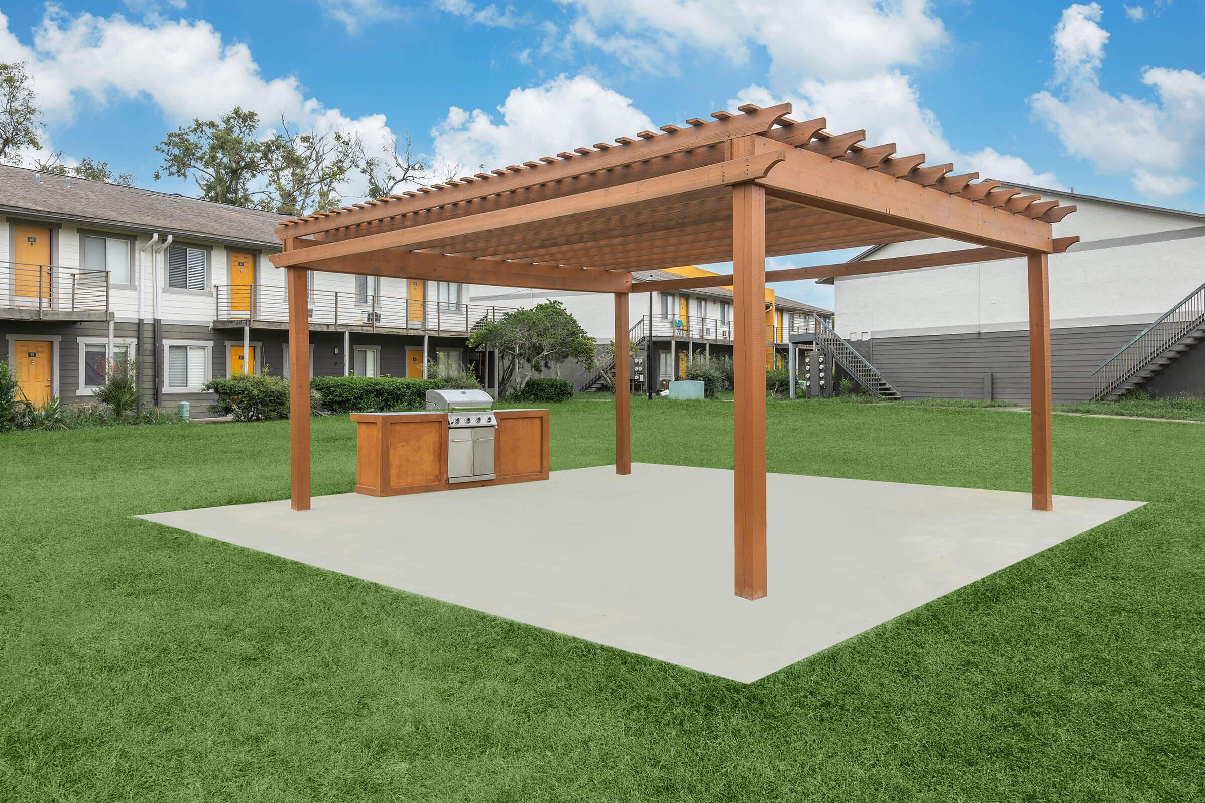 A wooden pergola stands over a concrete patio featuring a built-in outdoor grill. The setting is in a grassy courtyard surrounded by buildings with yellow doors and staircases, under a blue sky with fluffy clouds.