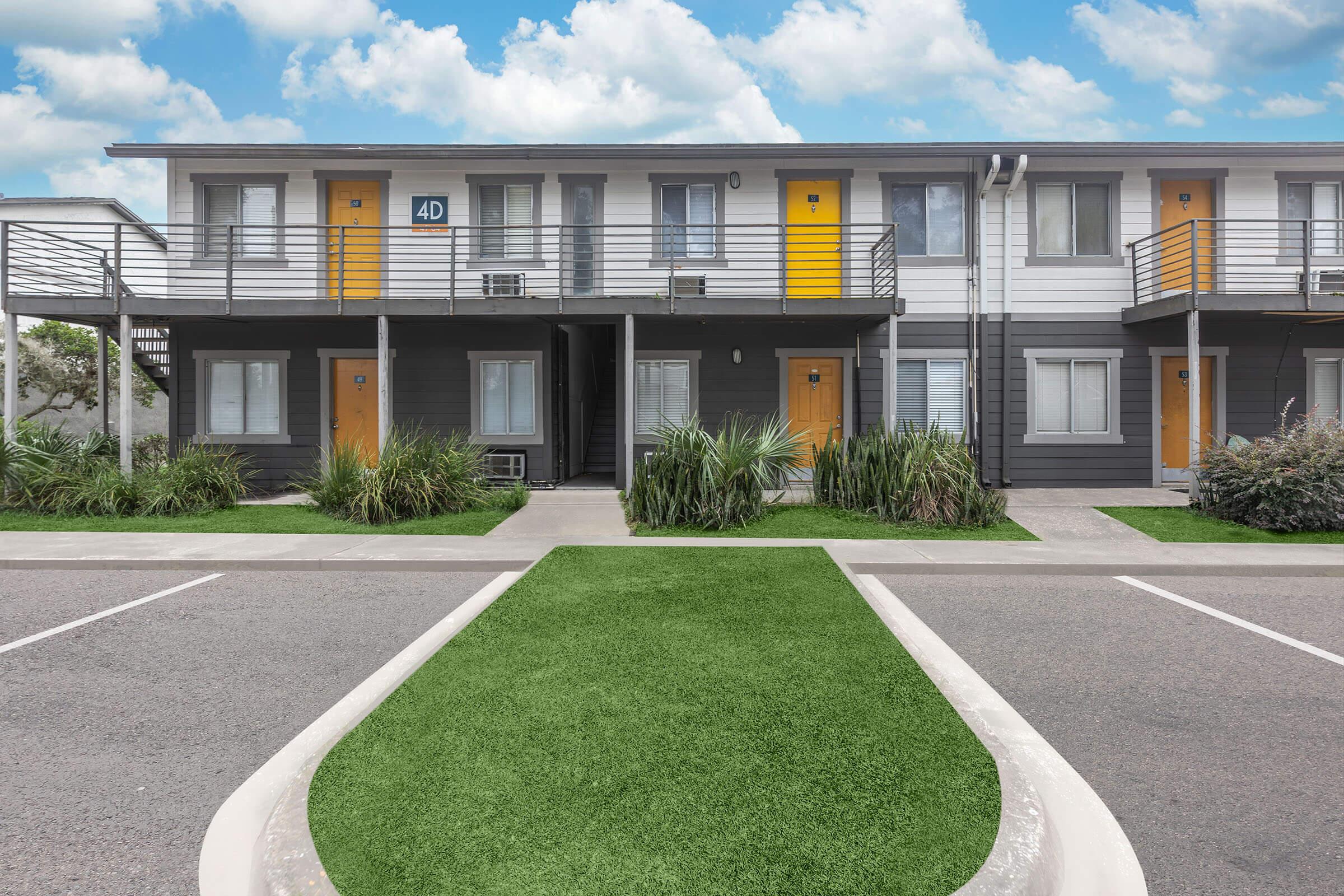 A modern two-story building featuring gray exteriors with yellow doors. The front area has well-maintained green grass and decorative plants. There are parking spaces visible in the foreground, and a clear blue sky with clouds in the background.