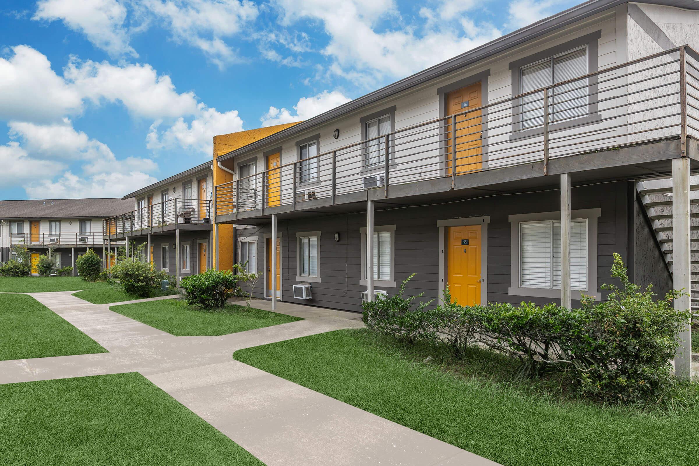 A row of modern two-story apartment buildings with gray siding and bright yellow doors. The path in front is lined with green lawns and shrubs, creating a welcoming outdoor space. The sky is partly cloudy, adding to the pleasant atmosphere of the residential area.