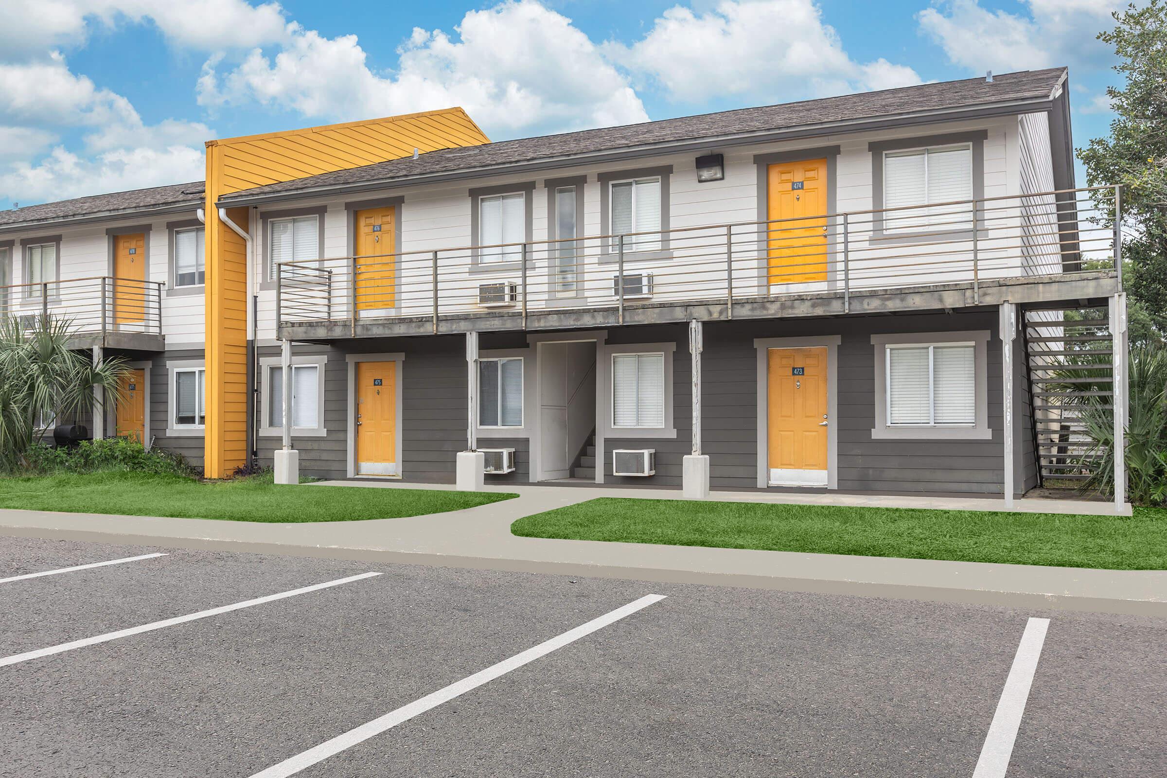 A two-story building with a modern design featuring gray and white exteriors, highlighted by bright orange doors. The structure has balconies and is surrounded by green grass. A parking lot is visible in the foreground, with a cloudy blue sky above.