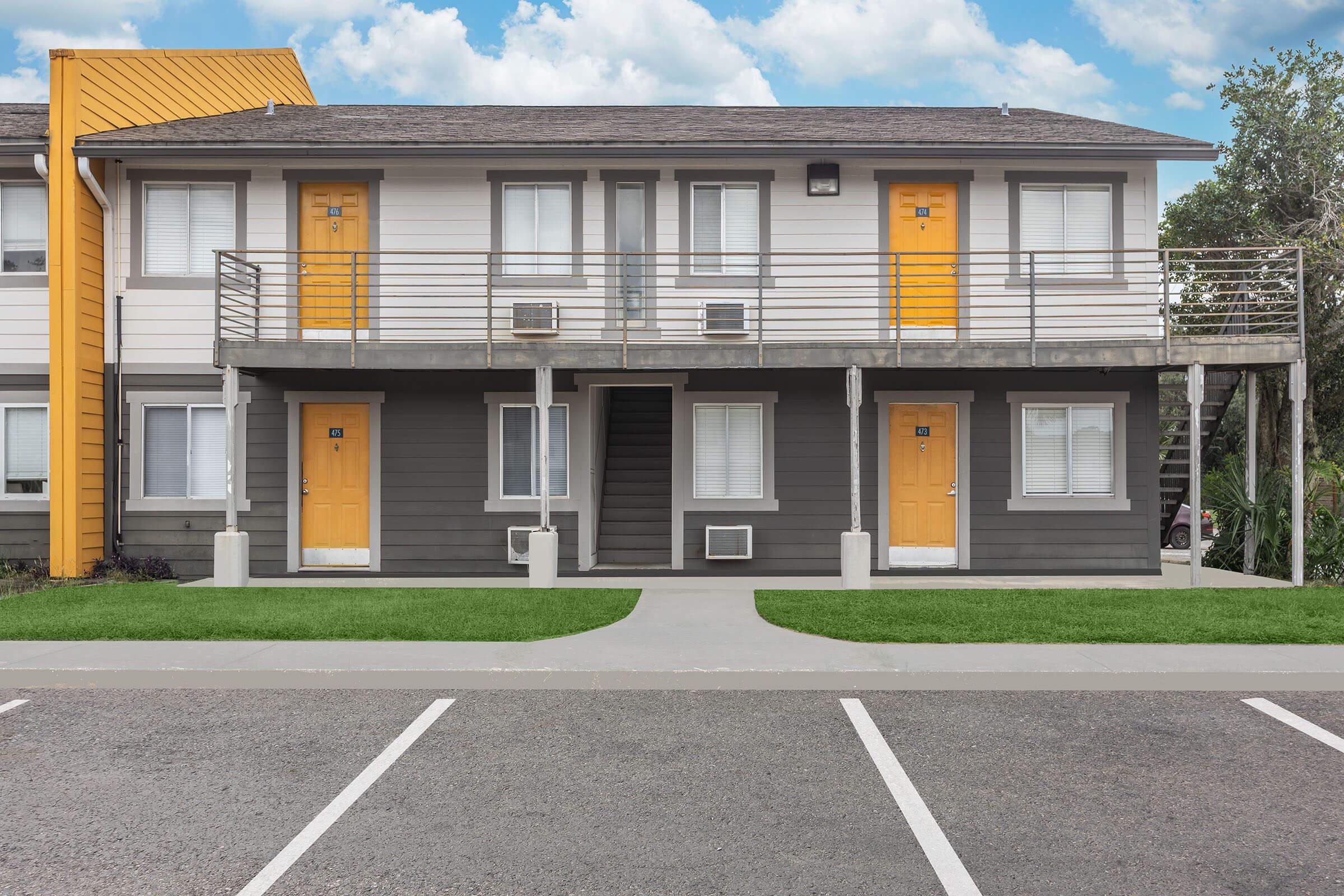 A two-story residential building featuring a modern design. The façade showcases a mix of gray and white siding with bright yellow doors. Each unit has a small balcony with railing. An asphalt parking lot is visible in front, with landscaped greenery surrounding the property and a blue sky above.