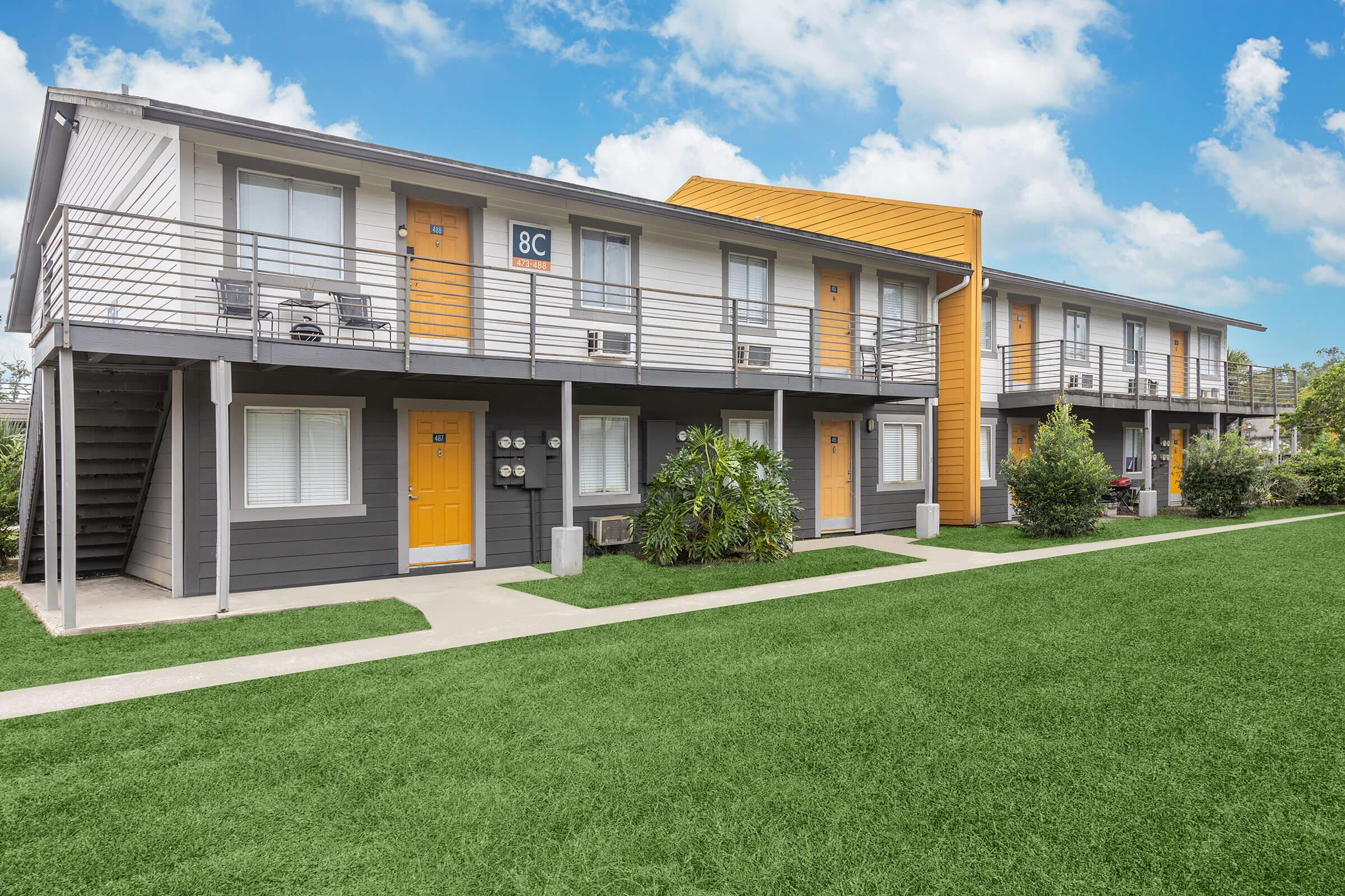 Two-story apartment building with a modern design, featuring yellow and gray exterior. The entrance doors are orange, and the path is lined with green grass. Lush plants are visible near the entrance, and the sky above is partly cloudy.