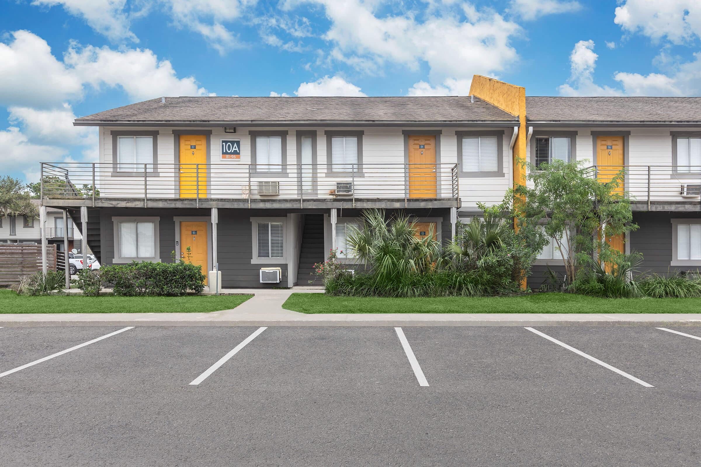 Exterior view of a two-story motel with orange doors and balconies. It features a well-maintained landscape, including shrubs and palm trees, with a clear blue sky in the background. The parking lot is visible in front, marked with white lines, indicating parking spaces.