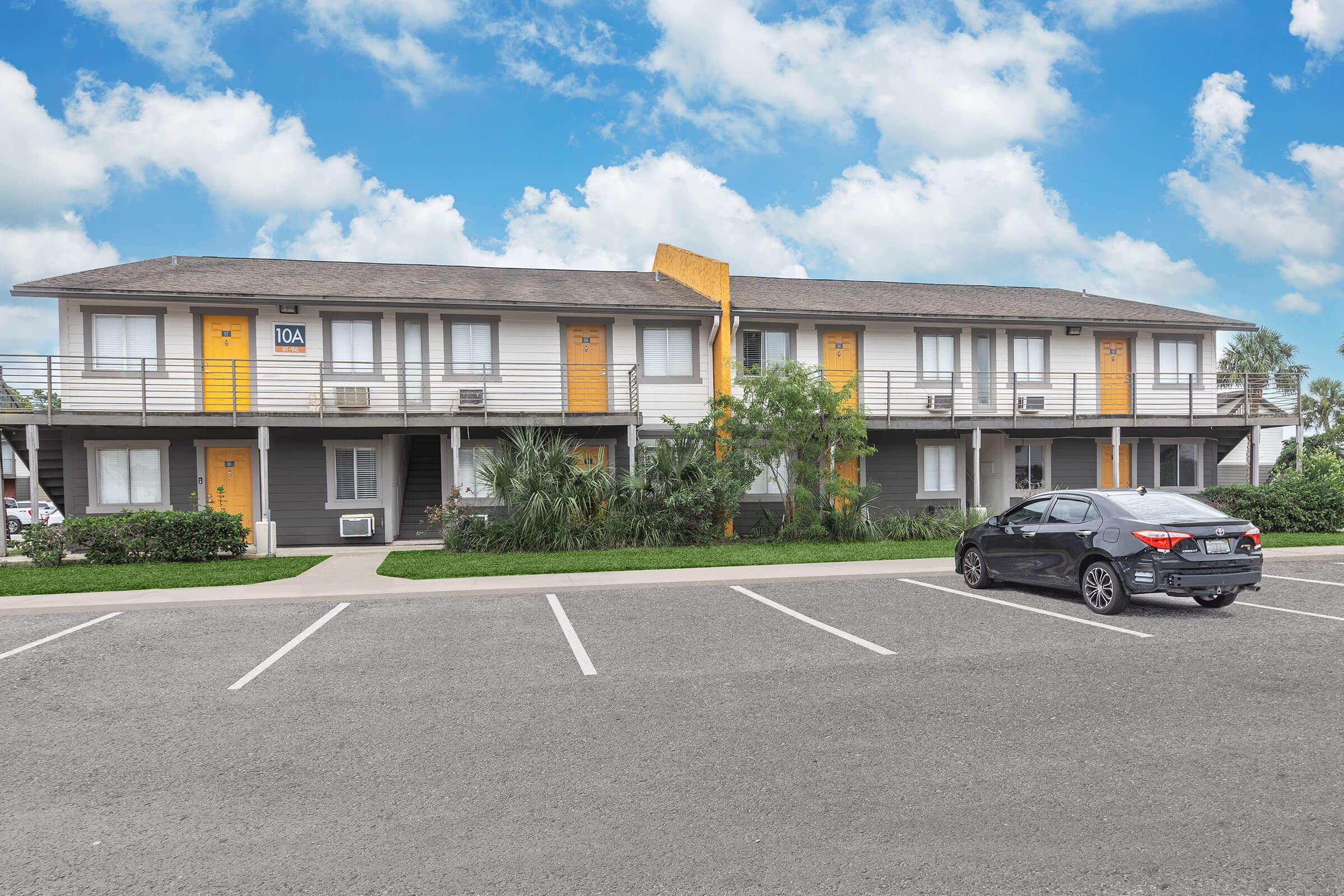 A two-story motel with yellow and gray exterior, featuring several rooms with balconies. Lush greenery surrounds the building, and a black car is parked in the foreground. The sky is bright blue with white clouds, creating a welcoming atmosphere.