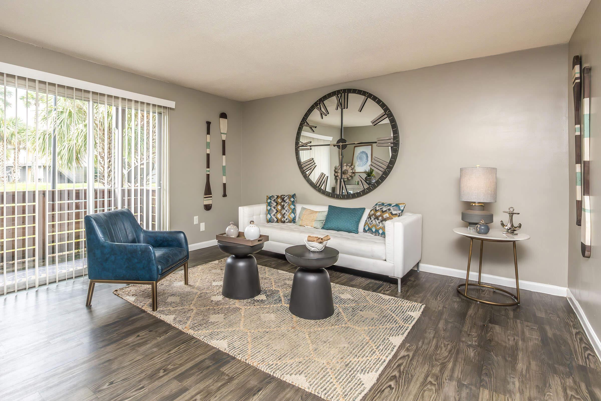 A modern living room featuring a white sofa with colorful pillows, a large round mirror on the wall, a blue accent chair, two black side tables, and a stylish lamp. The room has a warm color palette and a textured rug on wooden flooring, with sliding glass doors letting in natural light.