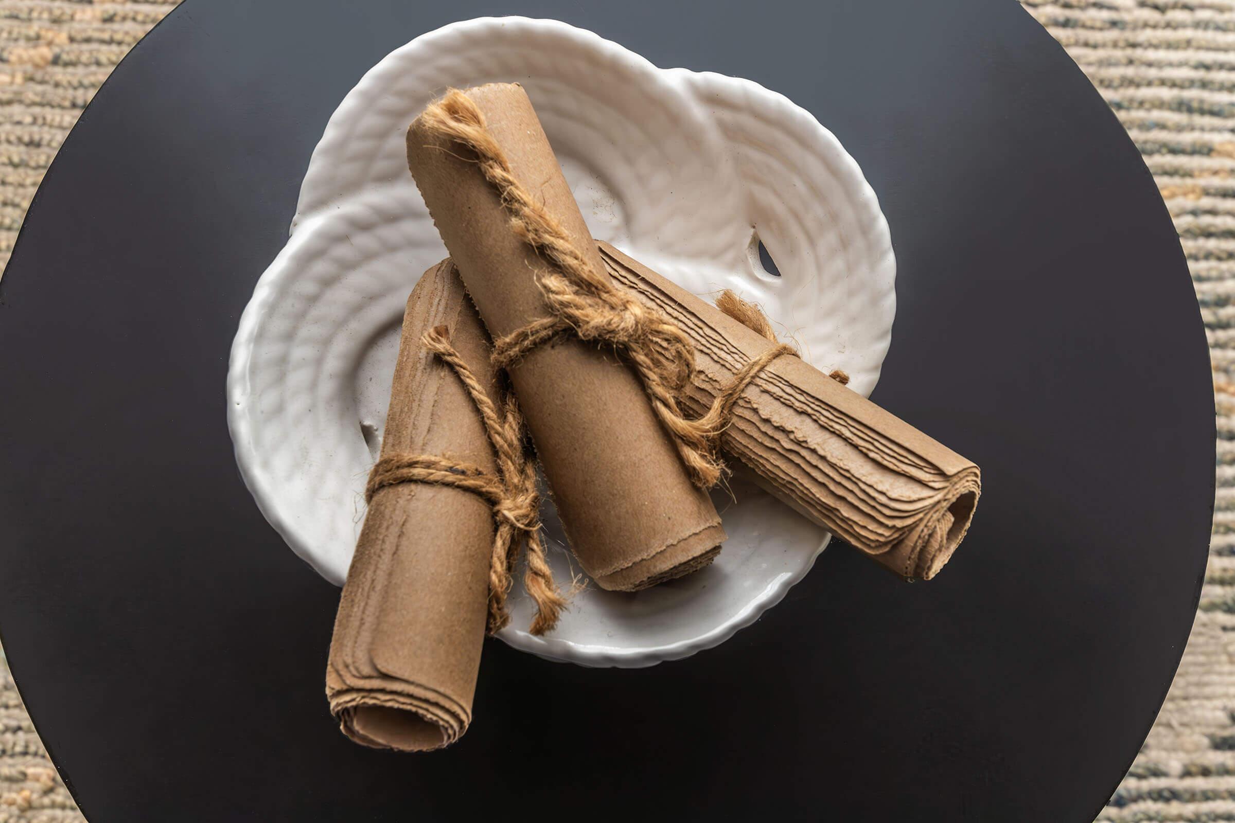 Three rolled pieces of paper tied with twine are placed in a decorative white bowl, sitting on a dark round table. The background features a textured, neutral-toned surface.