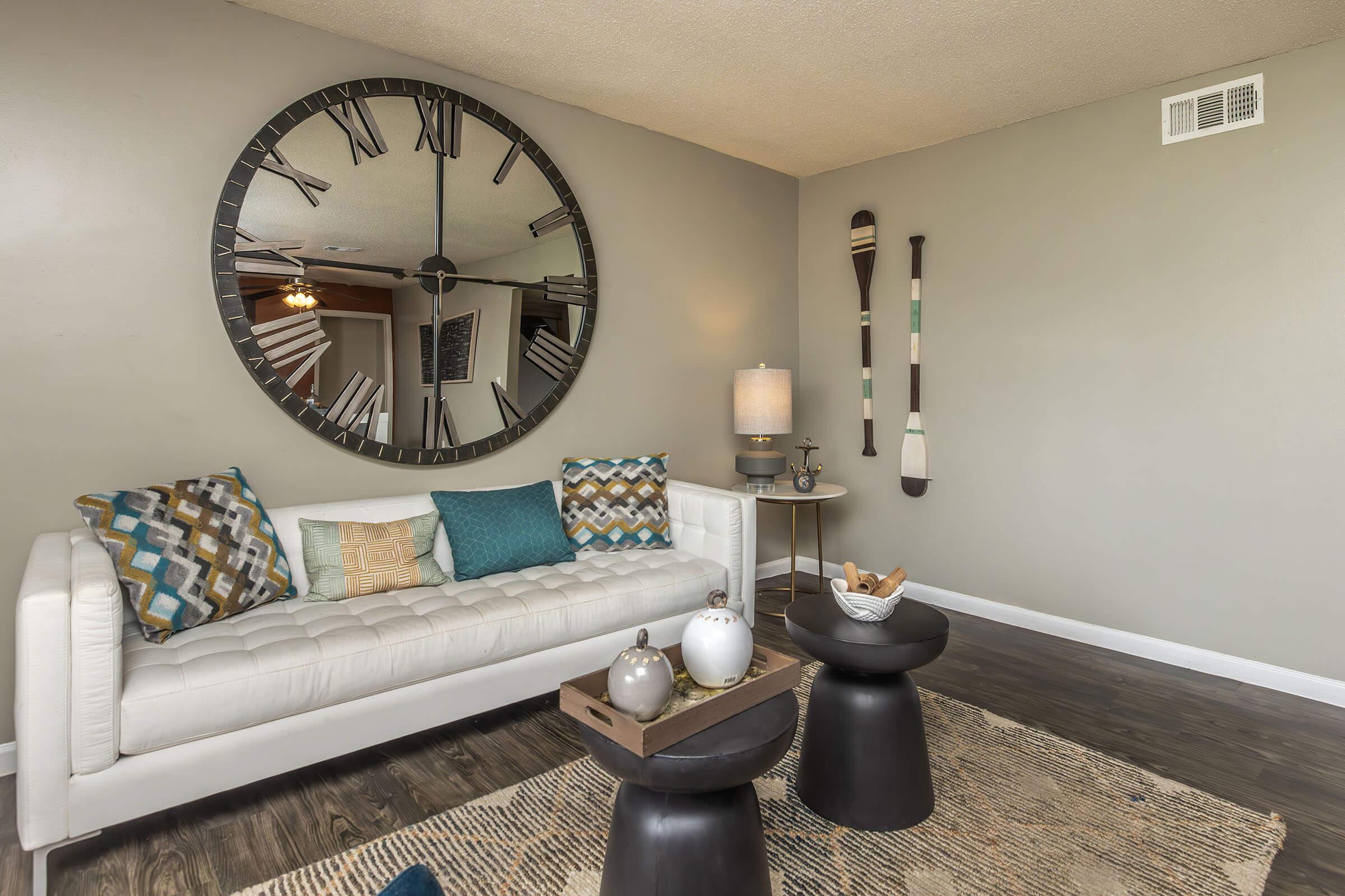 A modern living room featuring a white tufted sofa with decorative pillows, a large round wall clock, a small side table with a lamp, and a coffee table with decorative items. The walls are painted in a neutral tone, and there are two tall, artistic pieces hung on the wall. Natural light enhances the cozy ambiance.