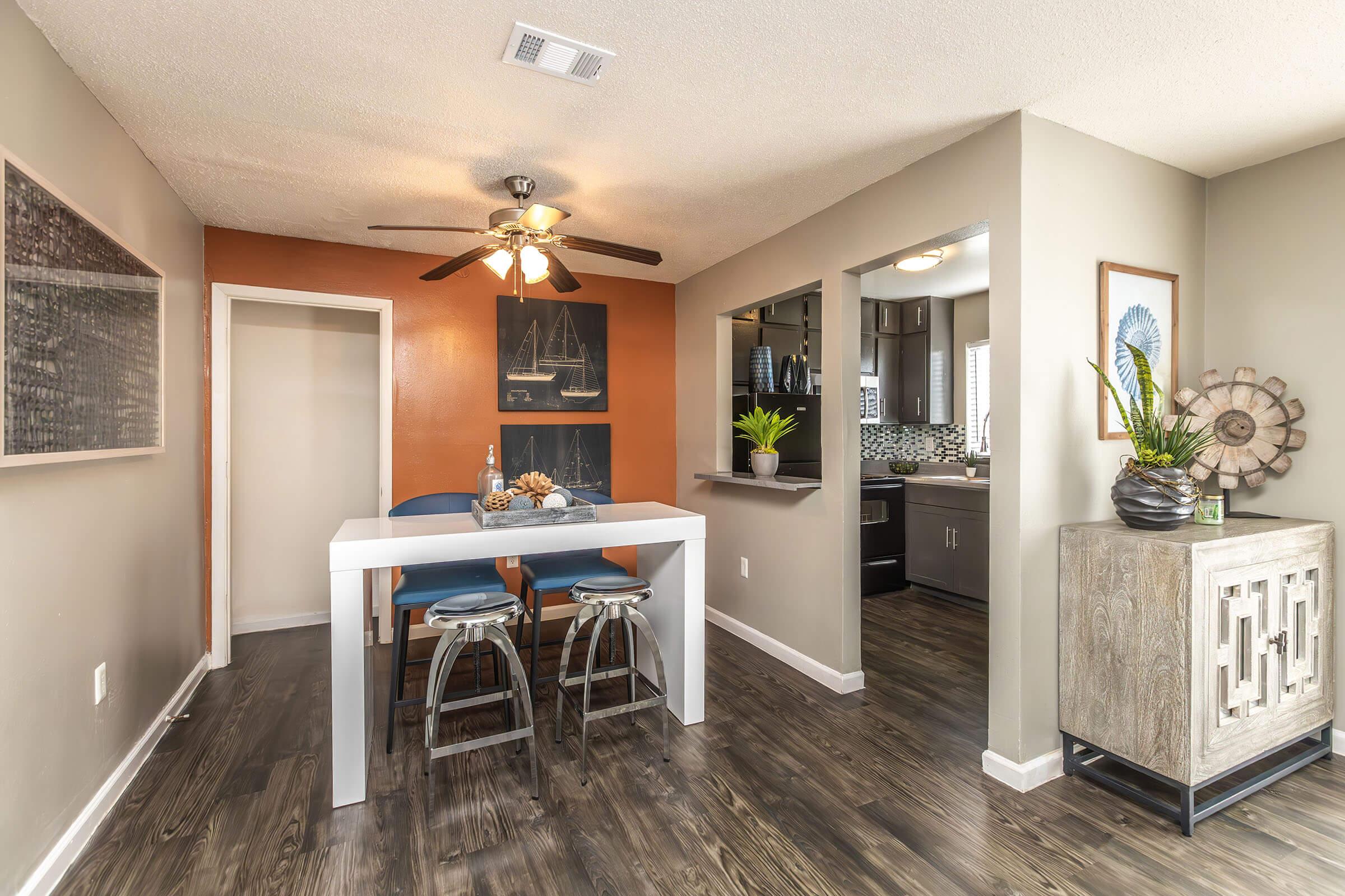 A cozy dining area featuring a white table with two blue stools, decorative items, and a ceiling fan. The walls are a mix of neutral tones with an orange accent, and there are framed artworks. A small bar area leads to a kitchen with modern appliances. The flooring is dark wood, adding warmth to the space.