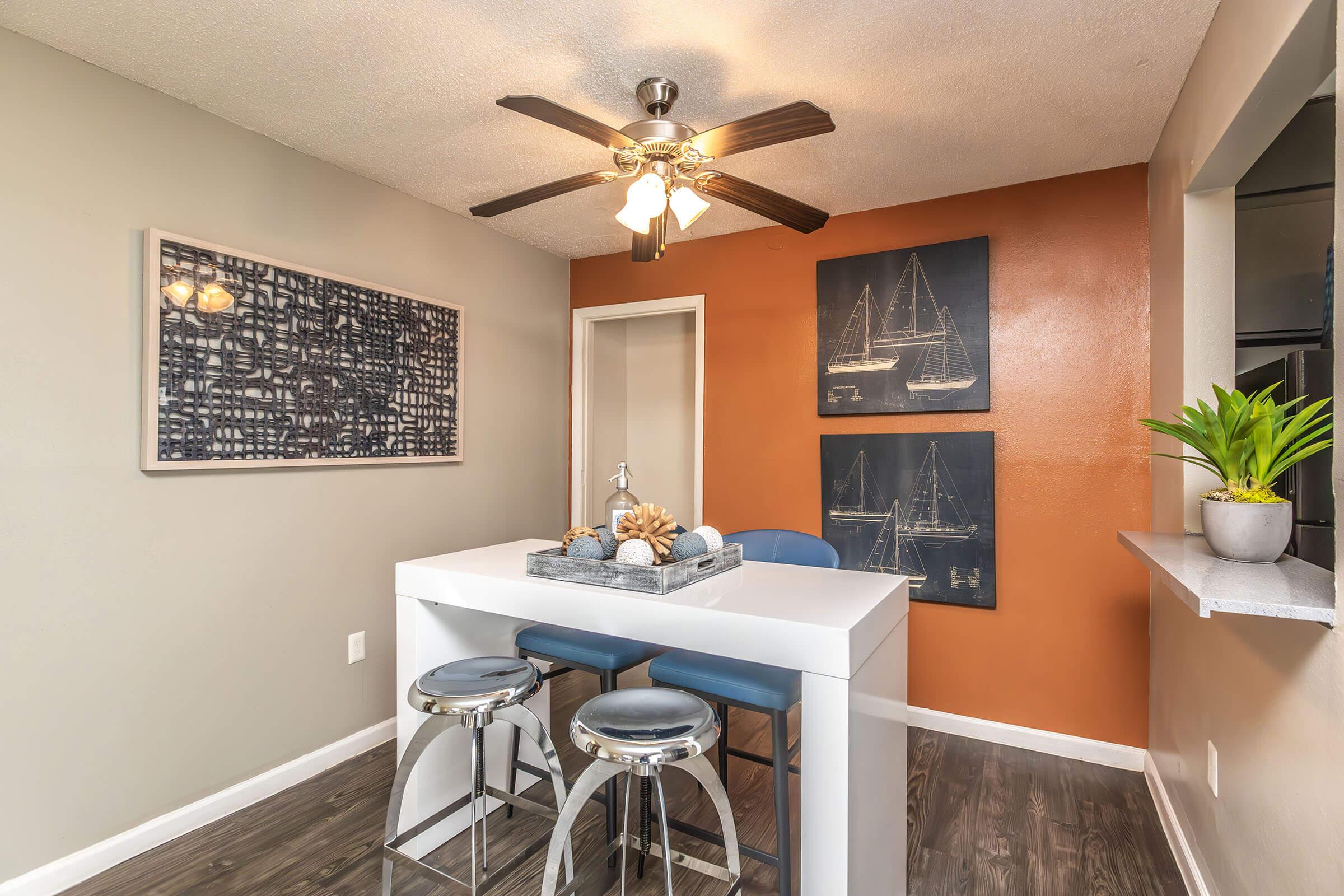 A modern dining area featuring a white table with decorative items, two blue upholstered bar stools, and a ceiling fan. The walls are a mix of light gray and a warm orange, with a framed abstract art piece and two framed nautical-themed images on the wall. A small potted plant adds a touch of greenery.