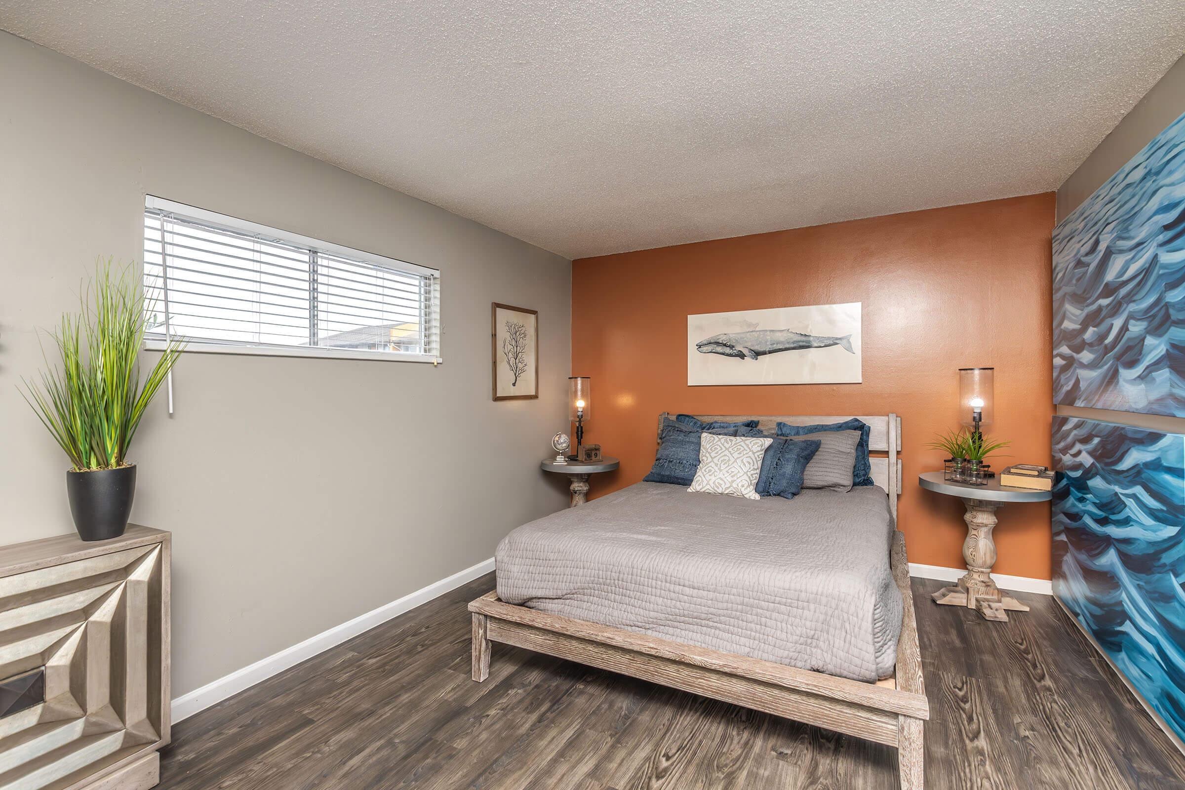 A cozy bedroom featuring a wooden bed with gray bedding, two bedside tables with lamps, and a wall art piece above the bed. One wall is painted orange, while the opposite wall has a blue wave design. A window lets in natural light and a potted plant adds a touch of greenery to the room. The flooring is wooden.