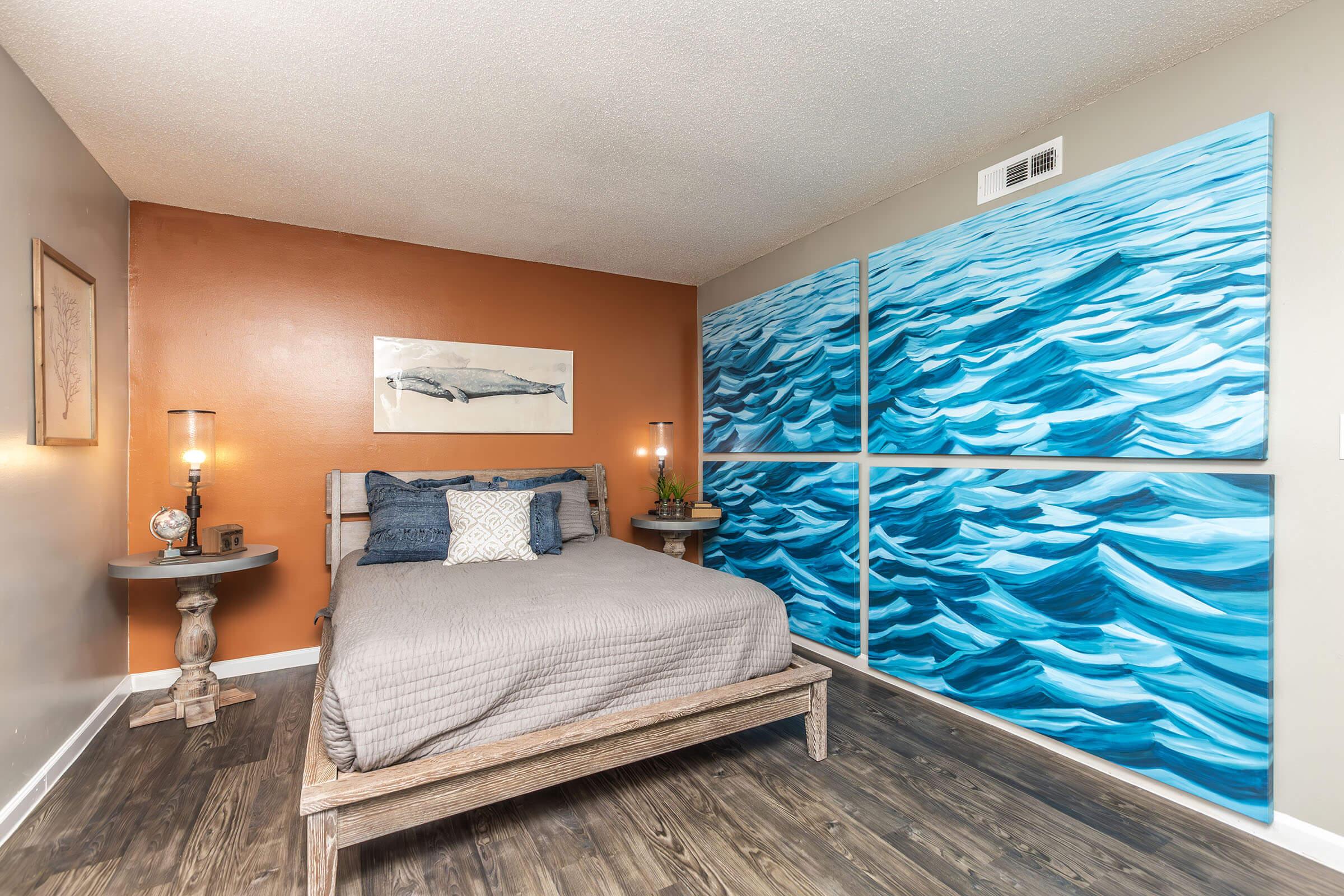 A modern bedroom featuring a wooden bedframe with a gray blanket and decorative pillows. The walls are painted in warm tones, with one accent wall in orange. There are two bedside tables, each with a lamp and decorative items. A large abstract artwork depicting waves is displayed prominently on the wall.