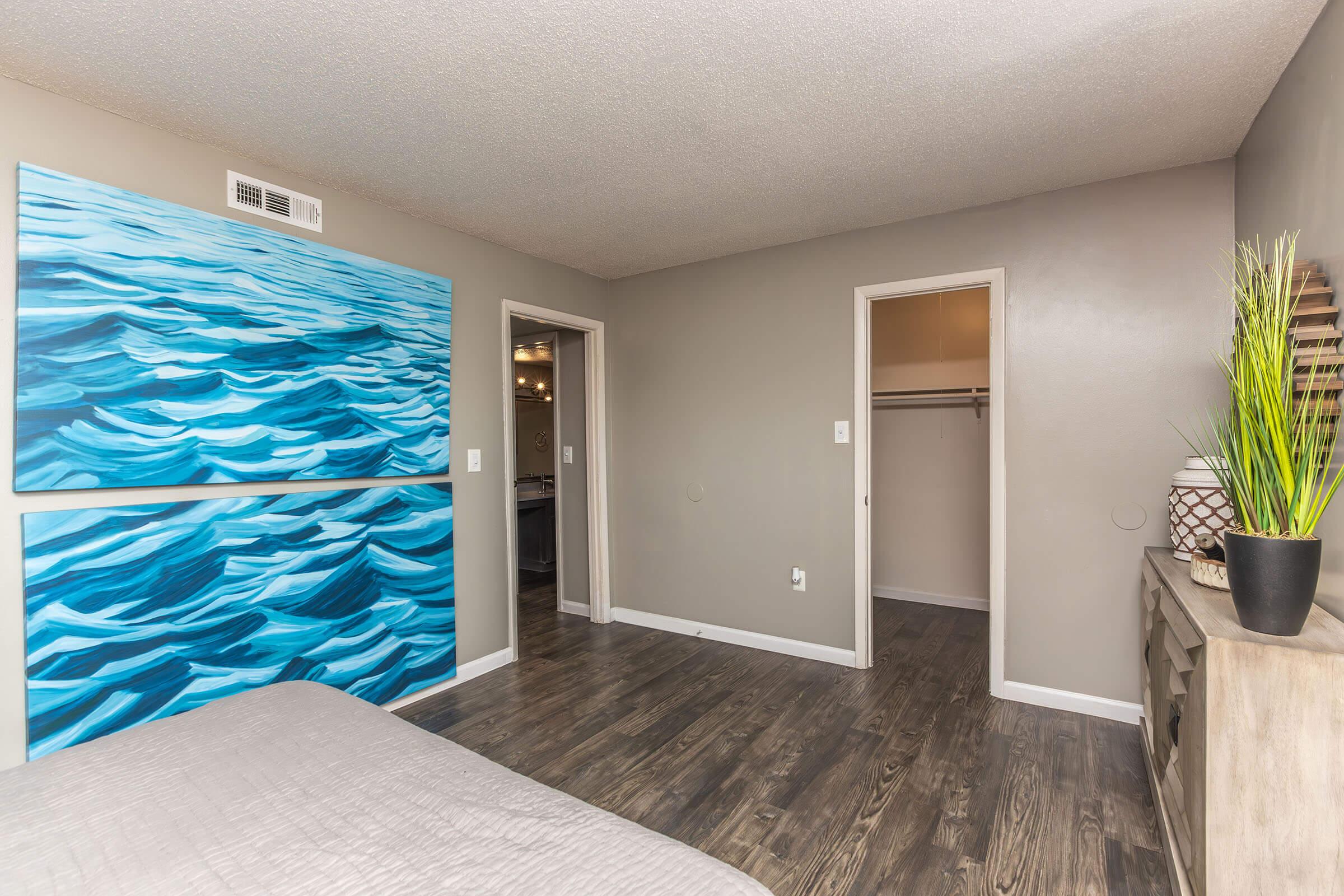 A modern bedroom featuring light gray walls, dark wood flooring, and a large abstract blue artwork. There are two doors: one leading to a closet and another possibly to a bathroom. A small decorative plant and a stylish unit with decorative items are also present in the room, creating a serene atmosphere.