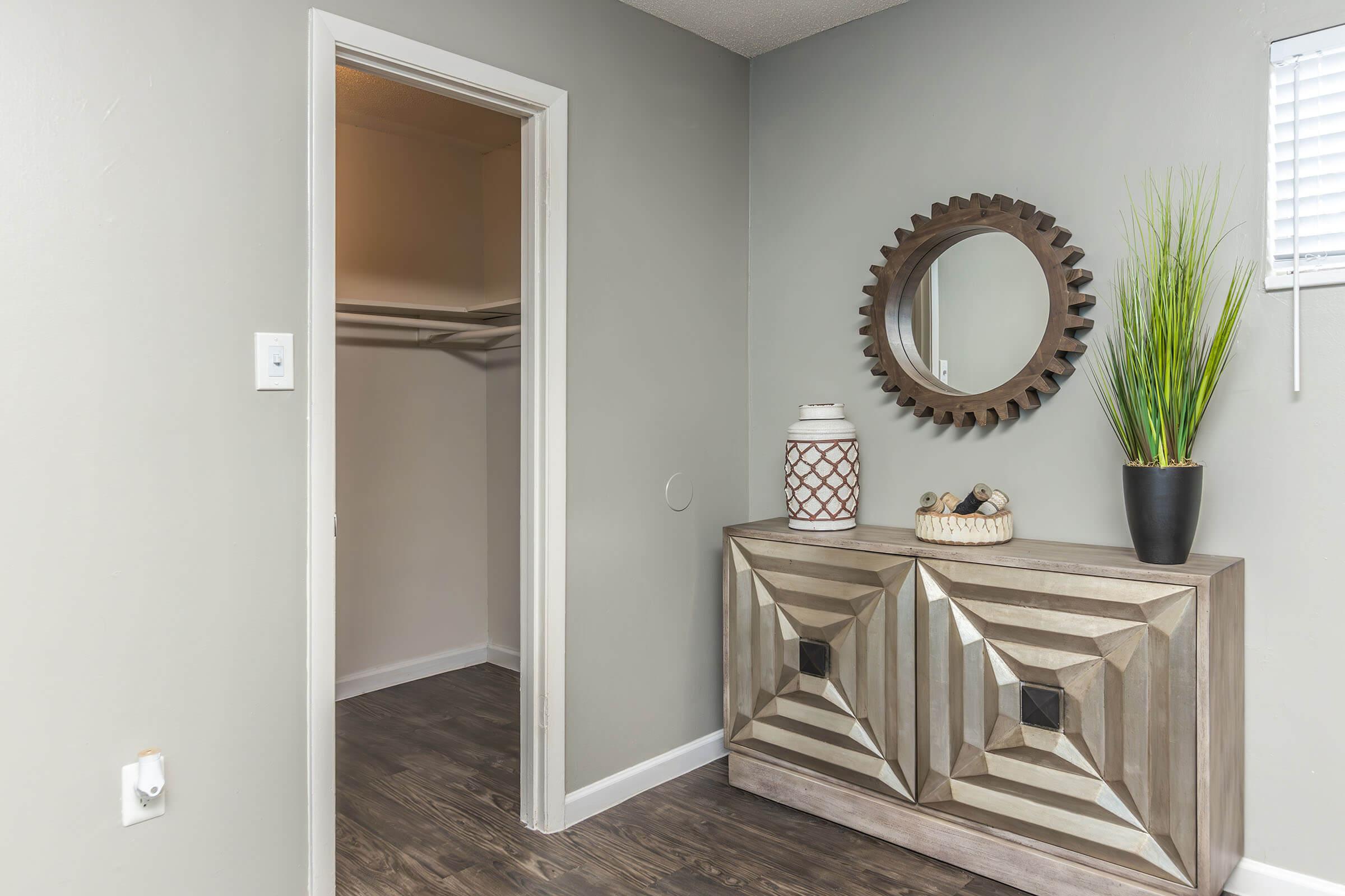 A small entryway features a textured wooden console table with decorative items on top, including a vase and a bowl. A round mirror hangs above the table, reflecting the room. To the left, an open door leads to a closet. The walls are painted in a soft gray, and a plant adds a touch of greenery.