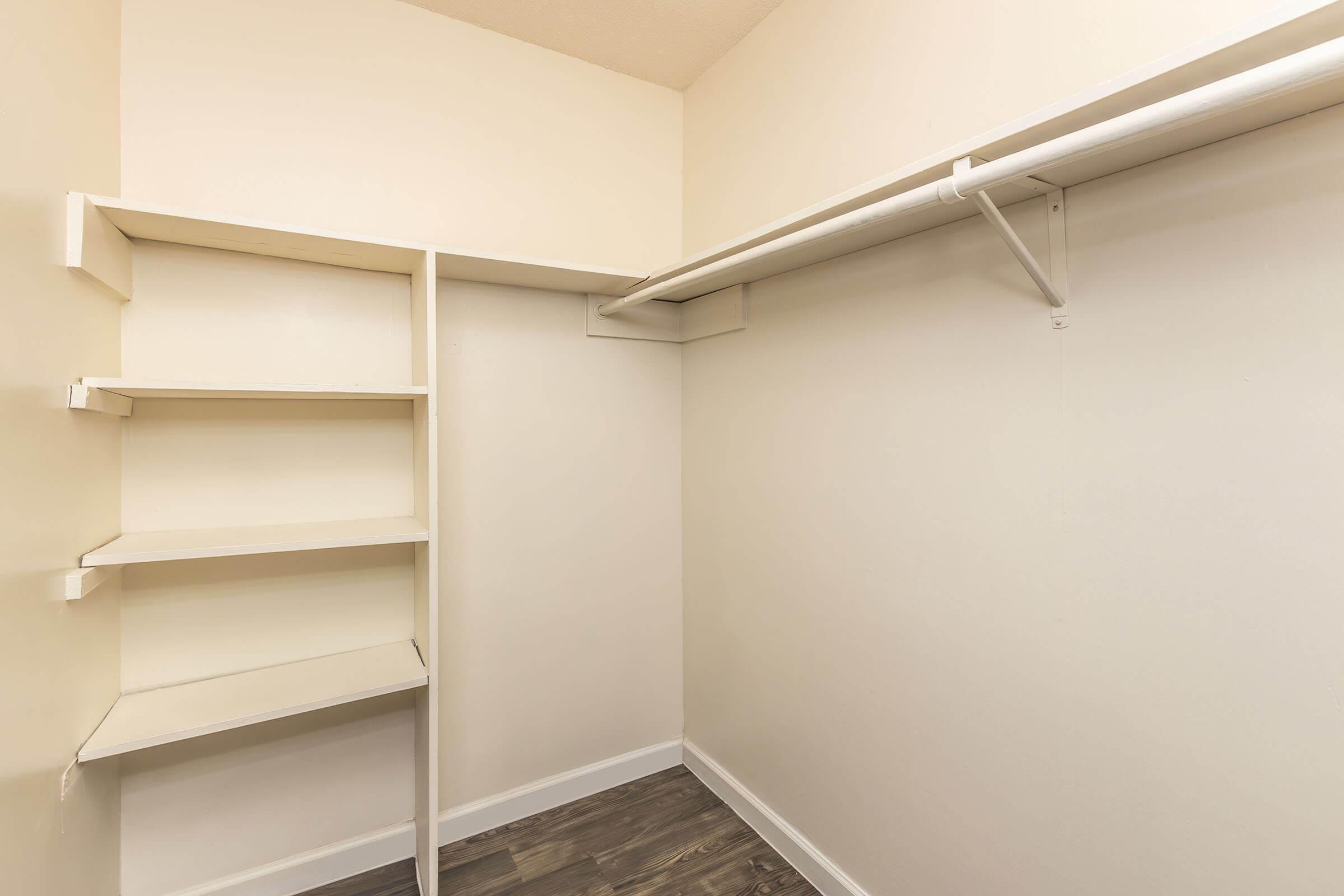 A well-organized empty closet featuring white walls and a light wooden floor. It includes built-in shelves on the left side and a hanging rod for clothing along the top. The spacious design allows for ample storage while maintaining a clean and minimalistic look.