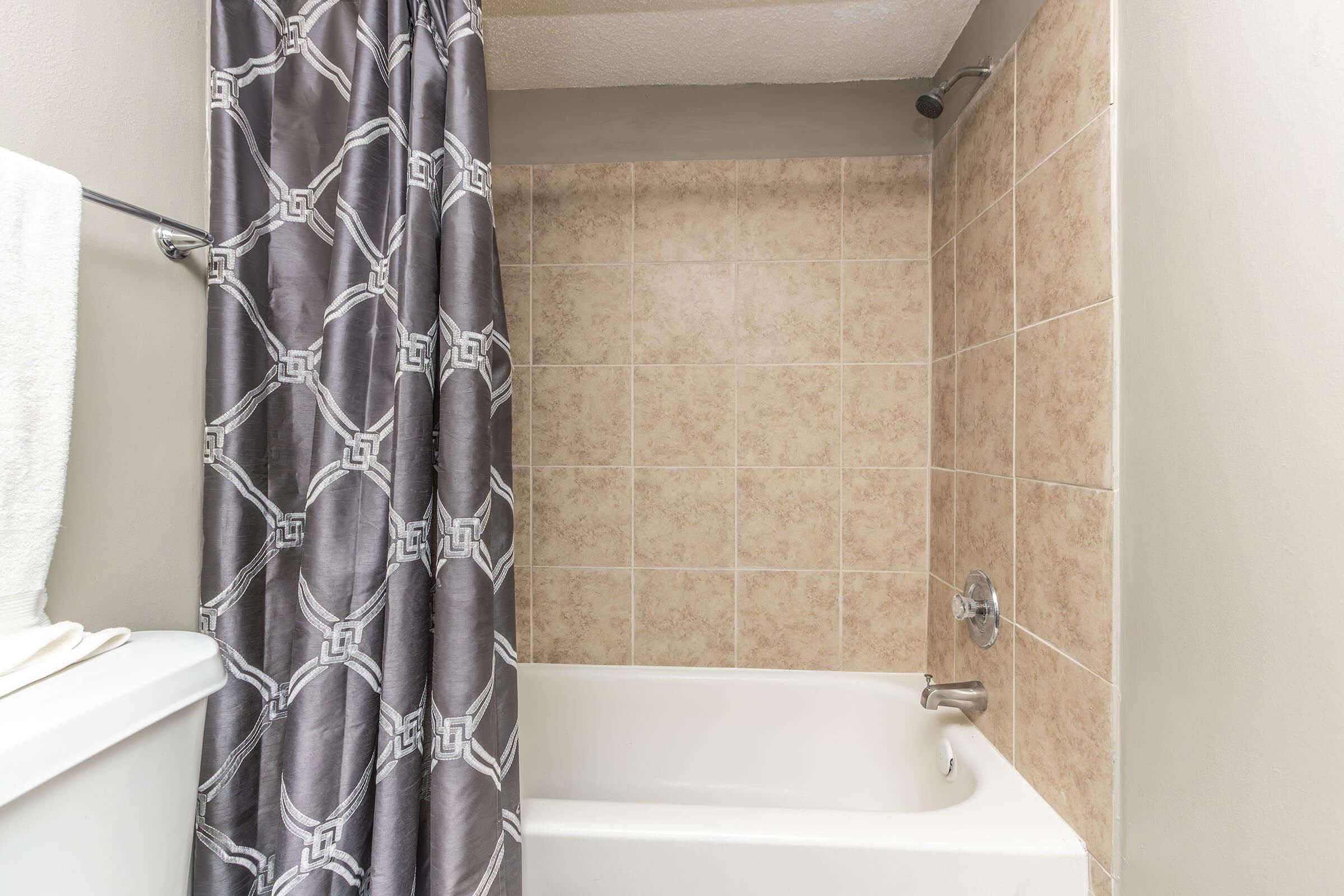 A bathroom scene featuring a white bathtub with a gray patterned shower curtain. The wall behind the tub is tiled in beige, while a towel is neatly hanging on a silver rack. The atmosphere is clean and simplistic, with muted colors creating a calm environment.