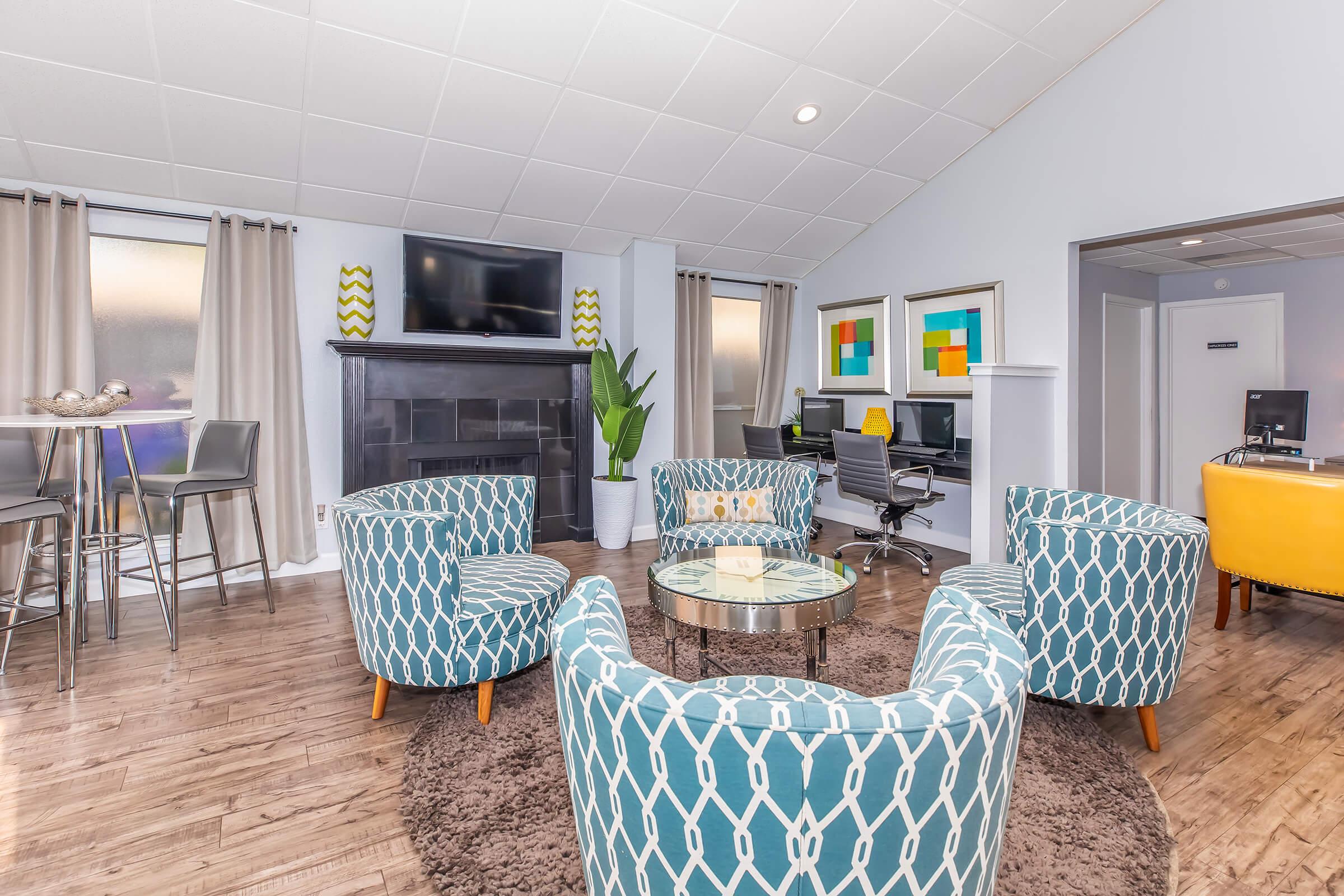 A modern communal living space featuring four teal patterned chairs arranged around a glass table. In the background, there are two computers at workstations, a flat-screen TV mounted on the wall, and large windows with sheer curtains. A large plant adds greenery, and there's a circular area rug on the wooden floor.