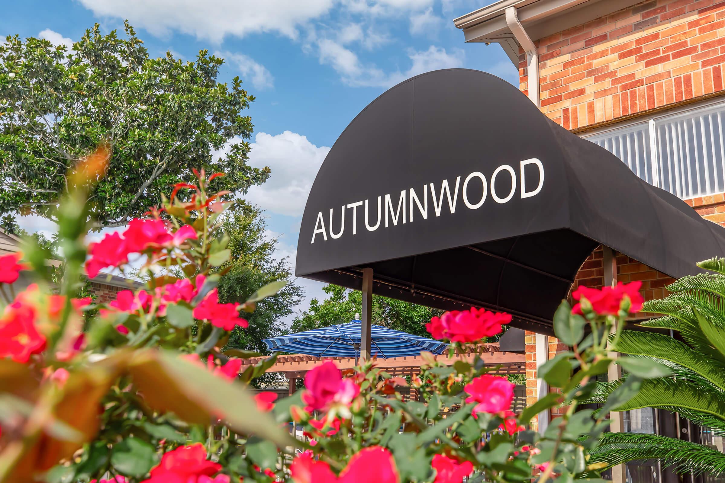 A close-up of a black awning displaying the word "AUTUMNWOOD," surrounded by vibrant pink flowers and greenery. The background features blue skies with scattered clouds, and the exterior of a brick building is partially visible.