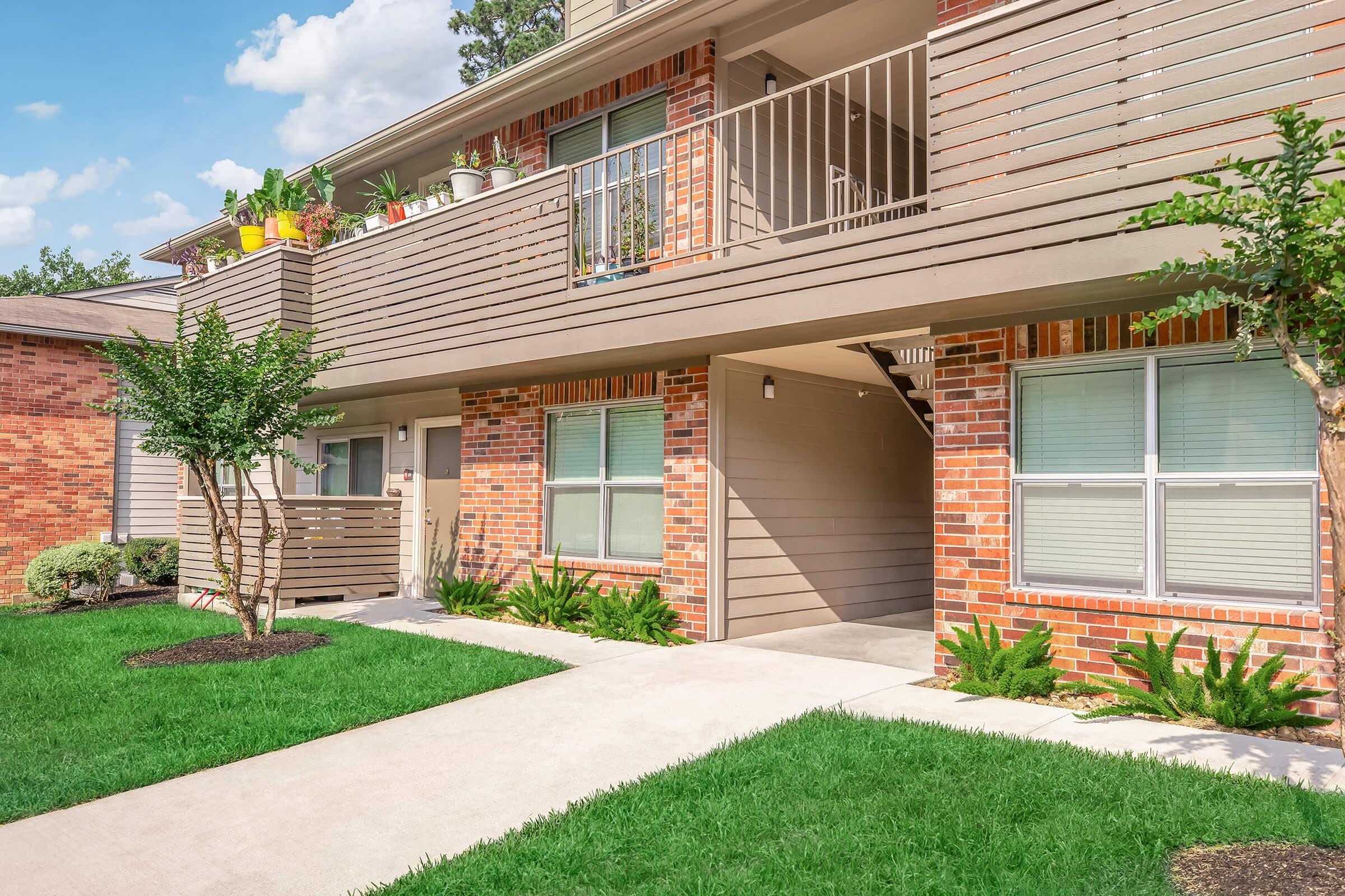 A well-maintained apartment complex featuring two balconies adorned with potted plants. The exterior is a mix of brick and siding, surrounded by lush green grass and small shrubs. Sidewalks lead up to the entrance, illuminated by soft natural light. Blue sky with scattered clouds is visible above.