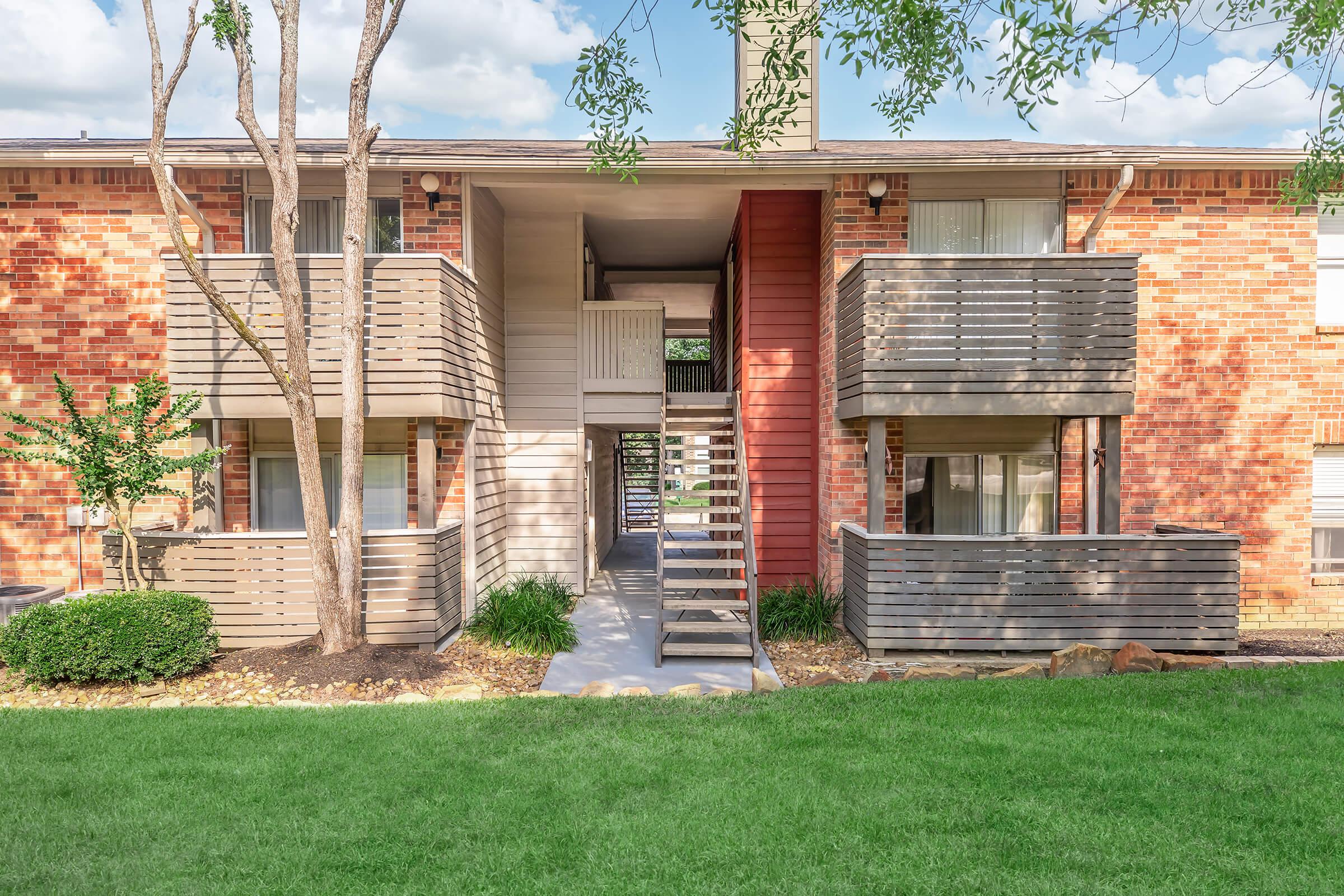 Two-story brick apartment building with wooden balconies. The building features a central staircase leading to the upper level, grassy lawns on either side, and trees providing some shade. The overall scene is bright and well-maintained, reflecting a welcoming residential environment.