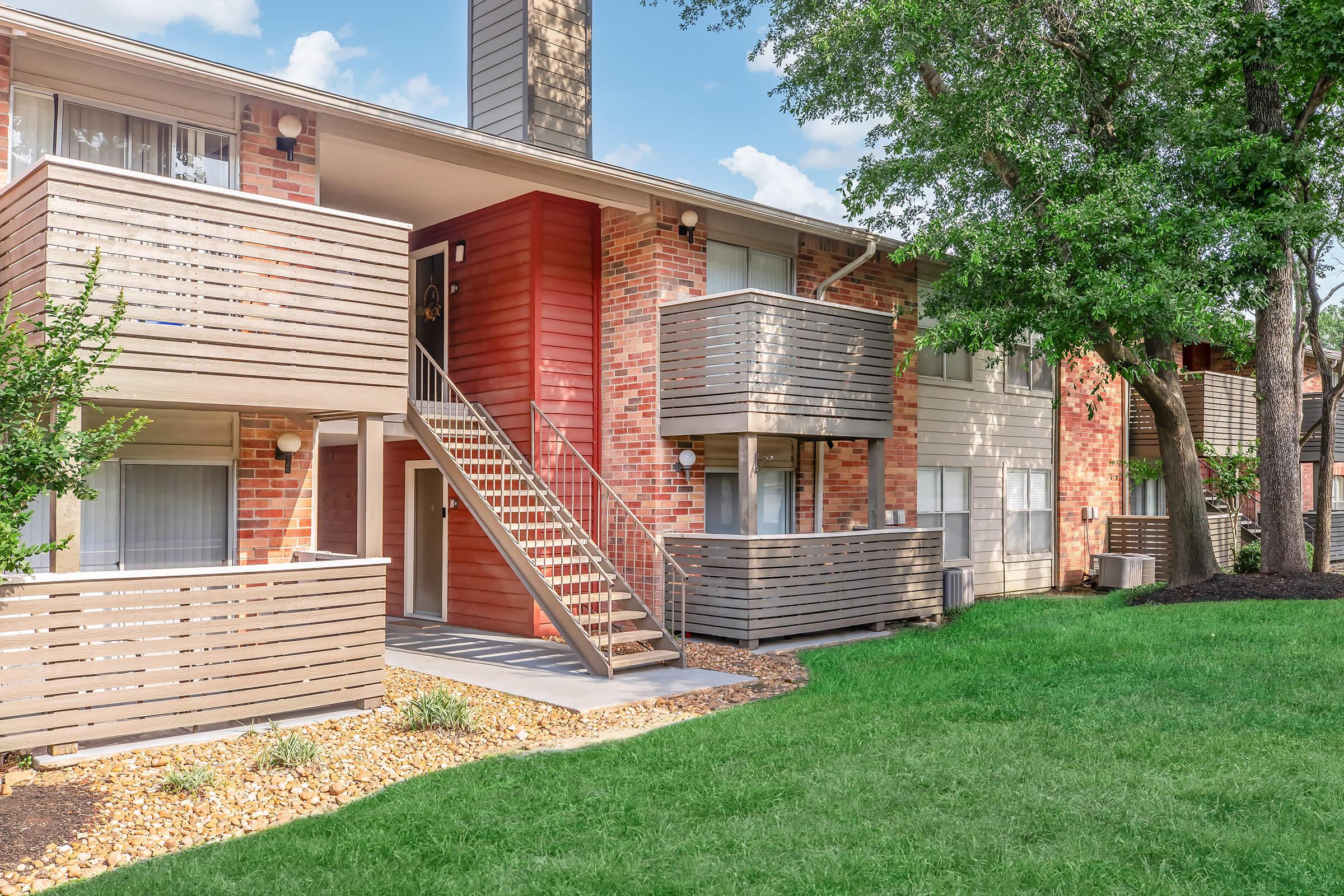 A multi-unit residential building featuring two levels of balconies, brick and wooden facade, and a well-maintained lawn with decorative stones and greenery. The image shows a staircase leading to the upper level and large windows allowing natural light into the apartments.