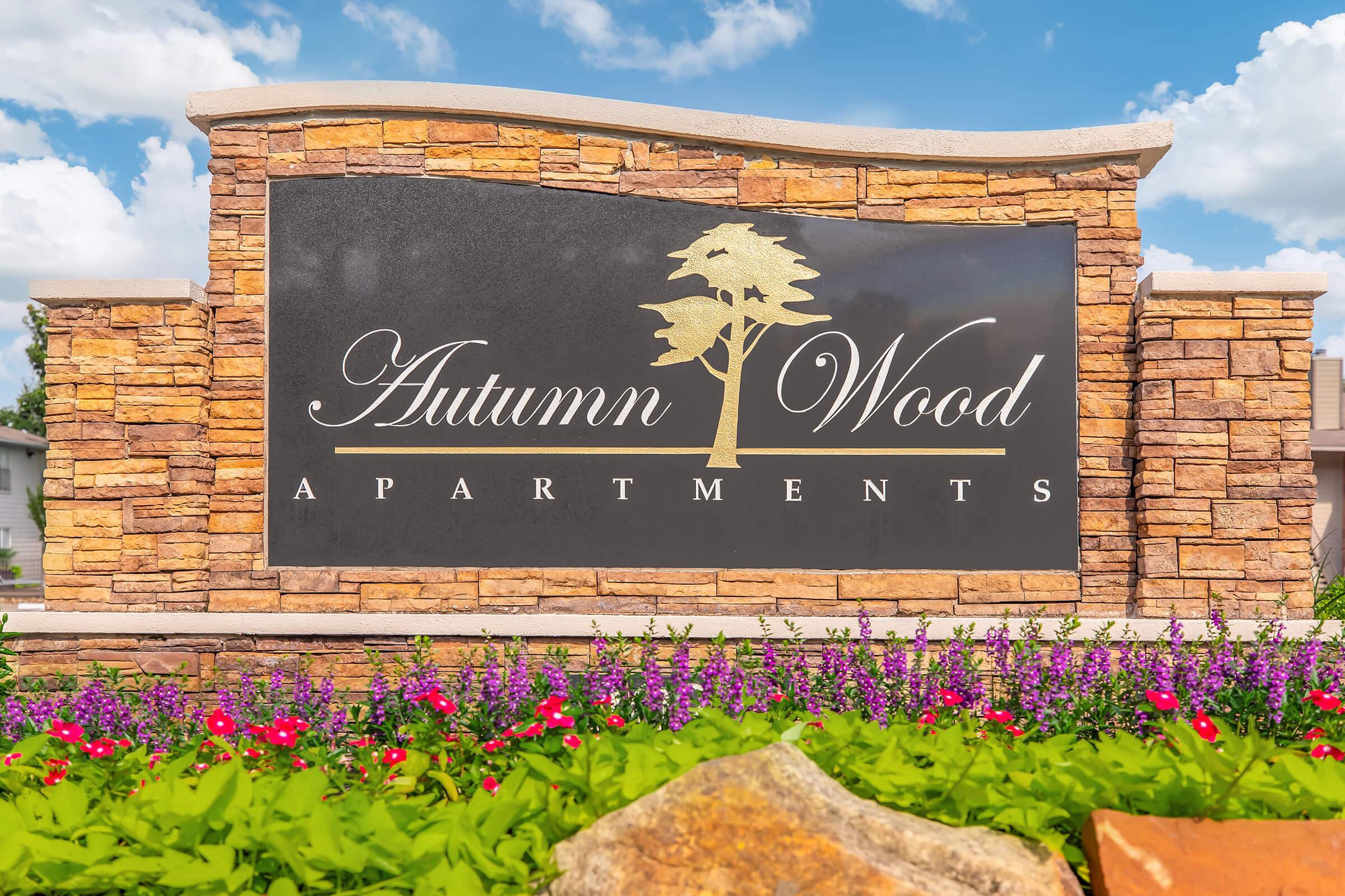 Sign for Autumn Wood Apartments with a stylized tree logo, surrounded by colorful flowers and greenery. The background features a blue sky with white clouds.