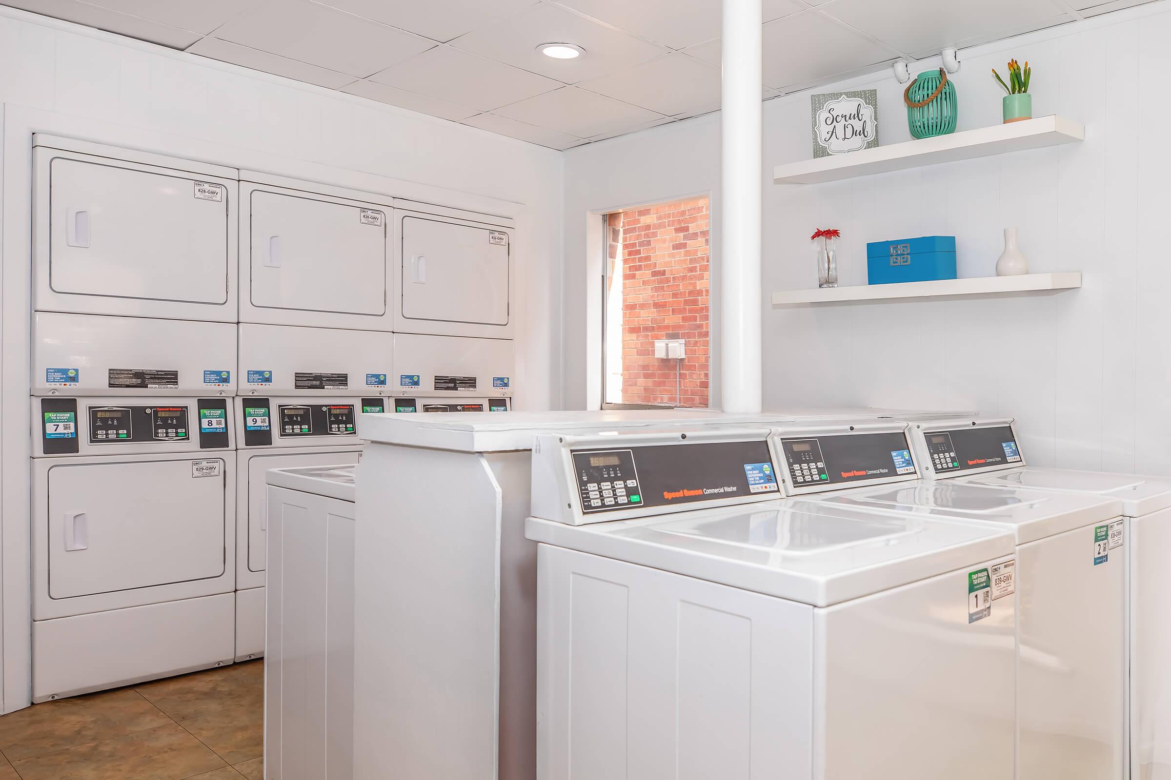 A modern laundry room featuring several stacked washing machines and dryers. The machines have digital control panels and are positioned against a white wall. There is a shelf above with decorative items, including a vase with flowers. Natural light enters through a window. The floor is tiled.