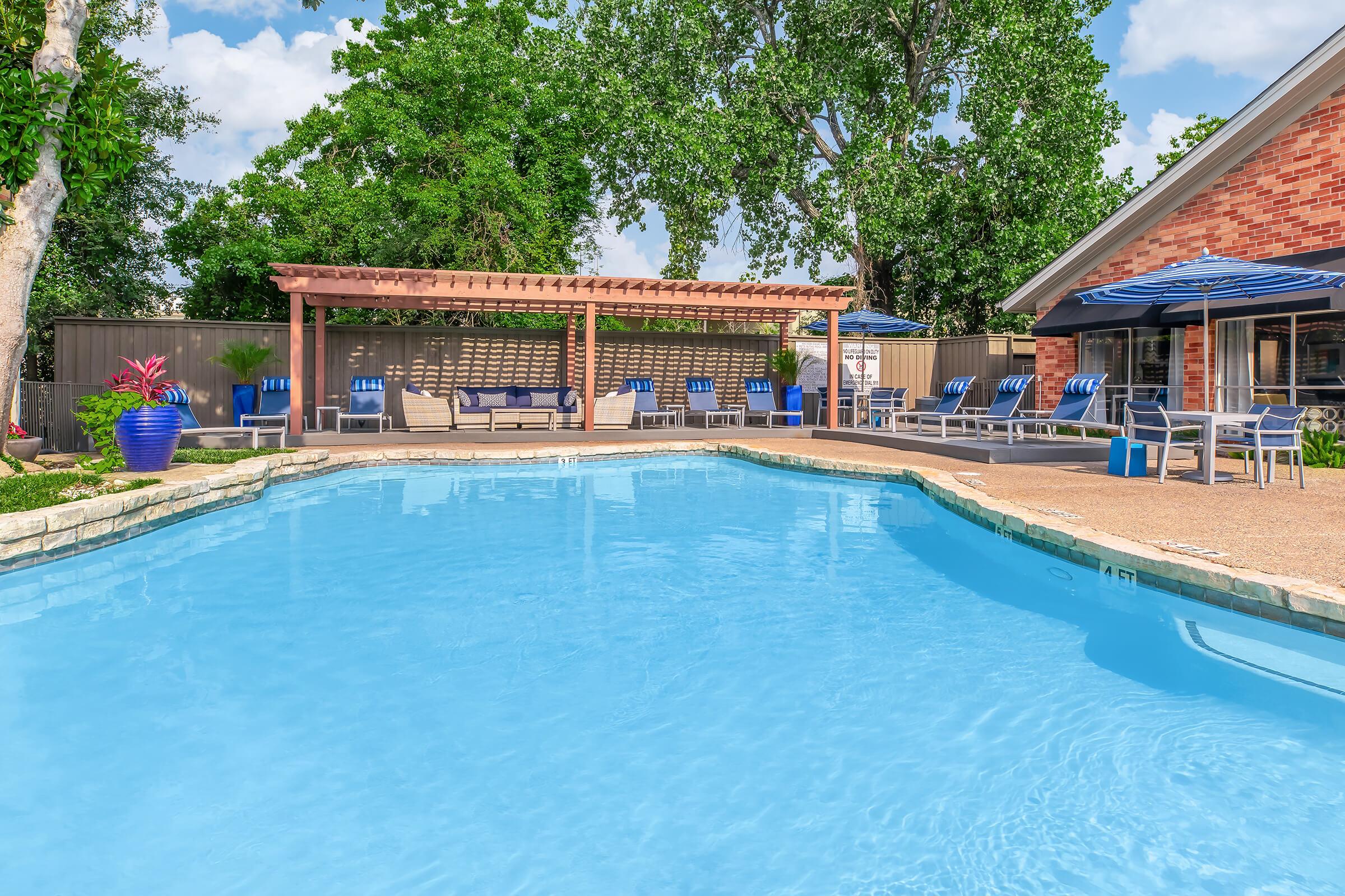 A sparkling blue swimming pool surrounded by a paved area and lounge chairs. A covered seating area with a wooden pergola is visible, along with blue umbrellas. Lush green trees and a brick wall provide a backdrop, completing the serene outdoor setting.