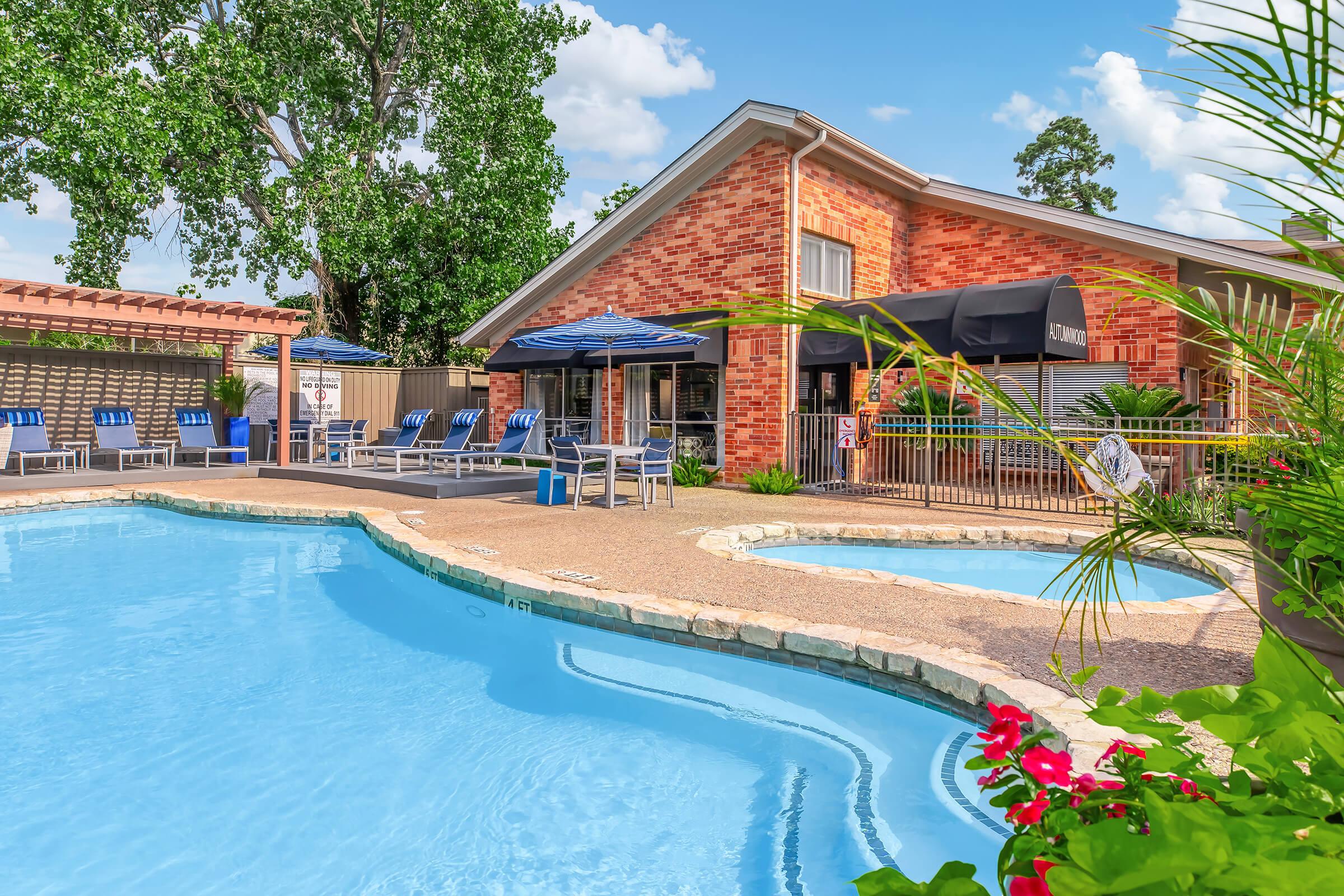 A bright outdoor pool area featuring clear blue water, surrounded by lounge chairs and umbrellas. In the background, a brick building with large windows and signage is visible, along with landscaped greenery and colorful flowers. A peaceful and inviting atmosphere for relaxation.