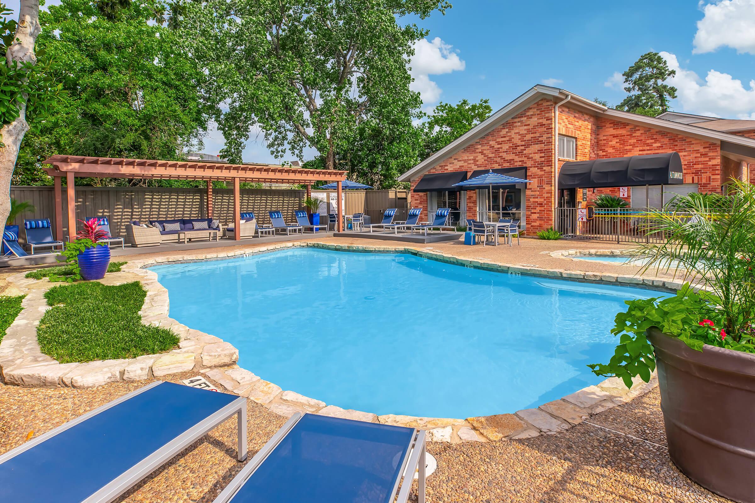 A sunny backyard featuring a clear blue swimming pool surrounded by lounge chairs and a stone deck. Nearby, there is a wooden pergola offering shade, and a brick house in the background with large windows. Lush greenery and colorful plants add a vibrant touch to the outdoor space.