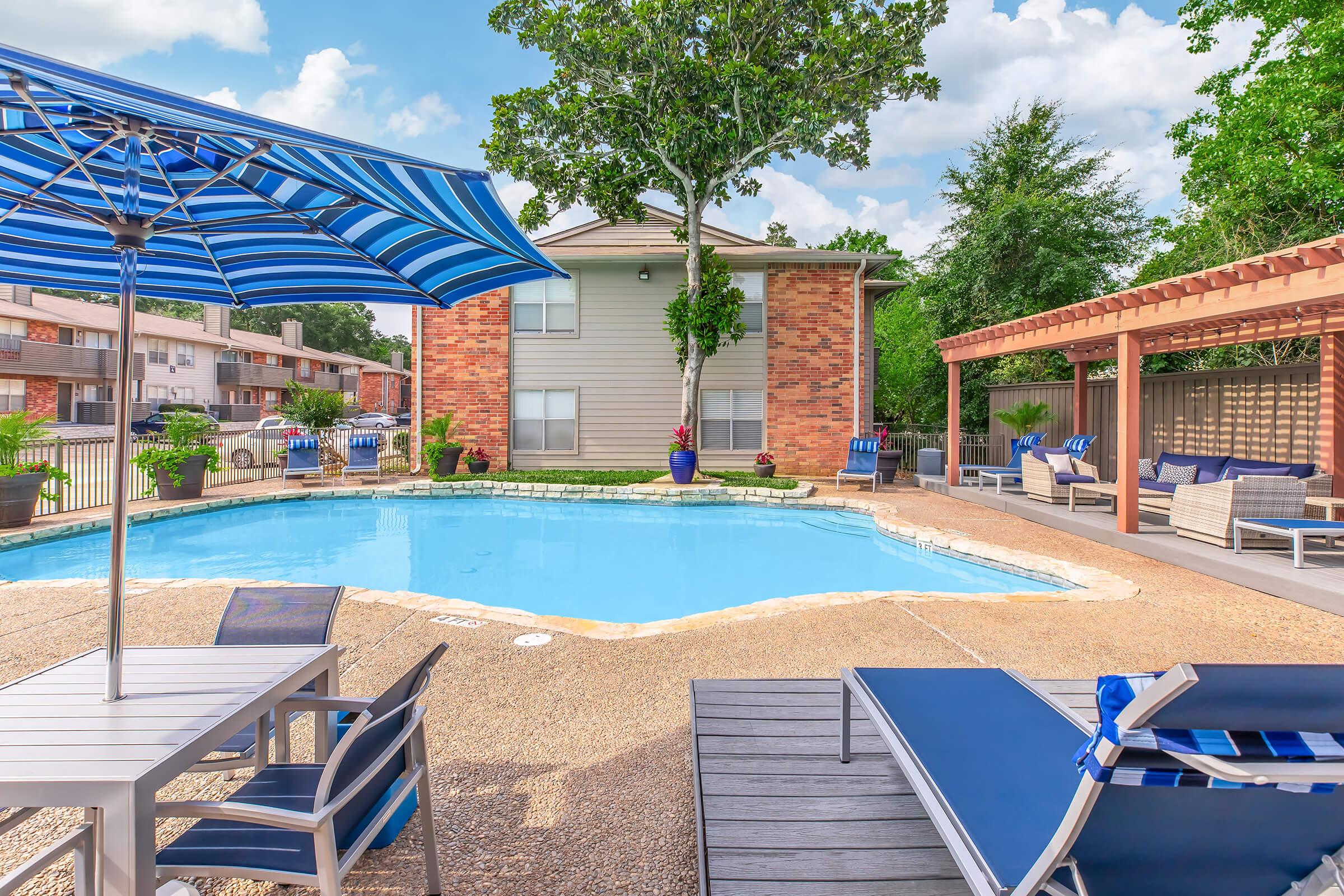 A sunny outdoor pool area featuring a blue and white striped umbrella, lounge chairs, and small tables. The pool is surrounded by landscaped greenery and a wooden pergola. In the background, there are two-story apartment buildings, creating a relaxed and inviting atmosphere.