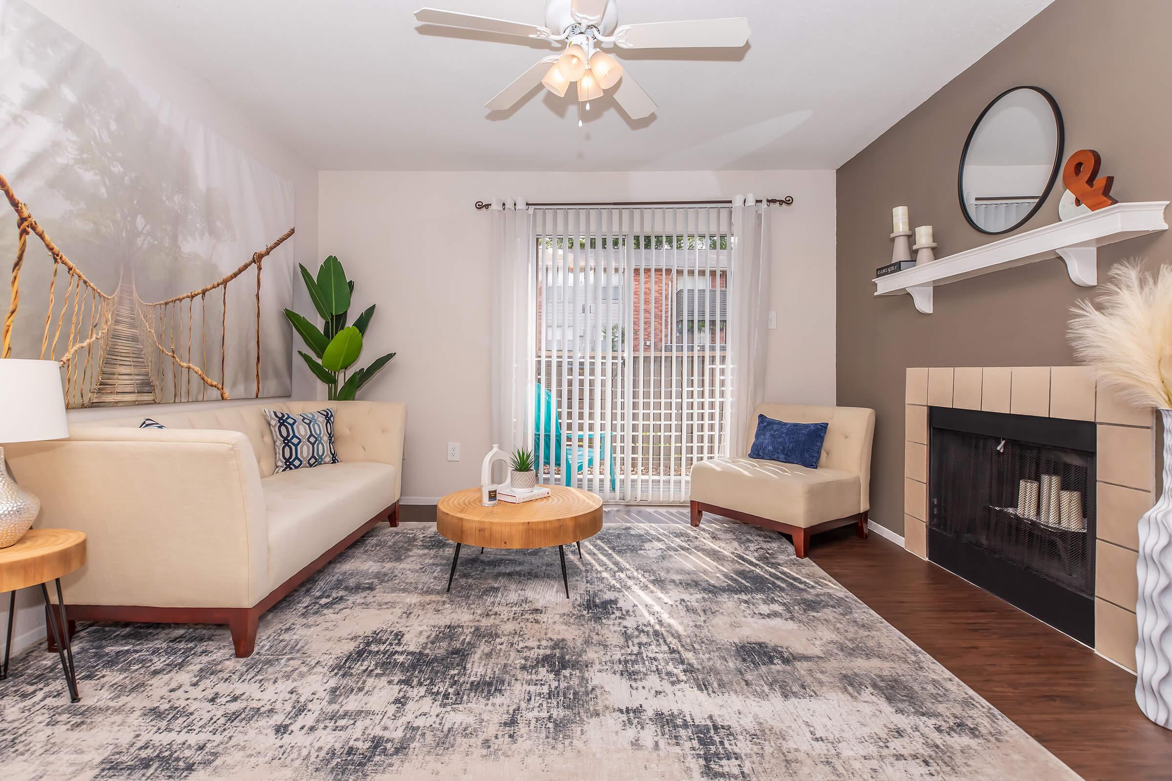 A modern living room featuring a light-colored sofa, a round wooden coffee table, and a large patterned area rug. A potted plant is in the corner, and a wall-mounted mirror reflects natural light. The room has a decorative fireplace and a sliding door leading to an outdoor area, creating a cozy atmosphere.