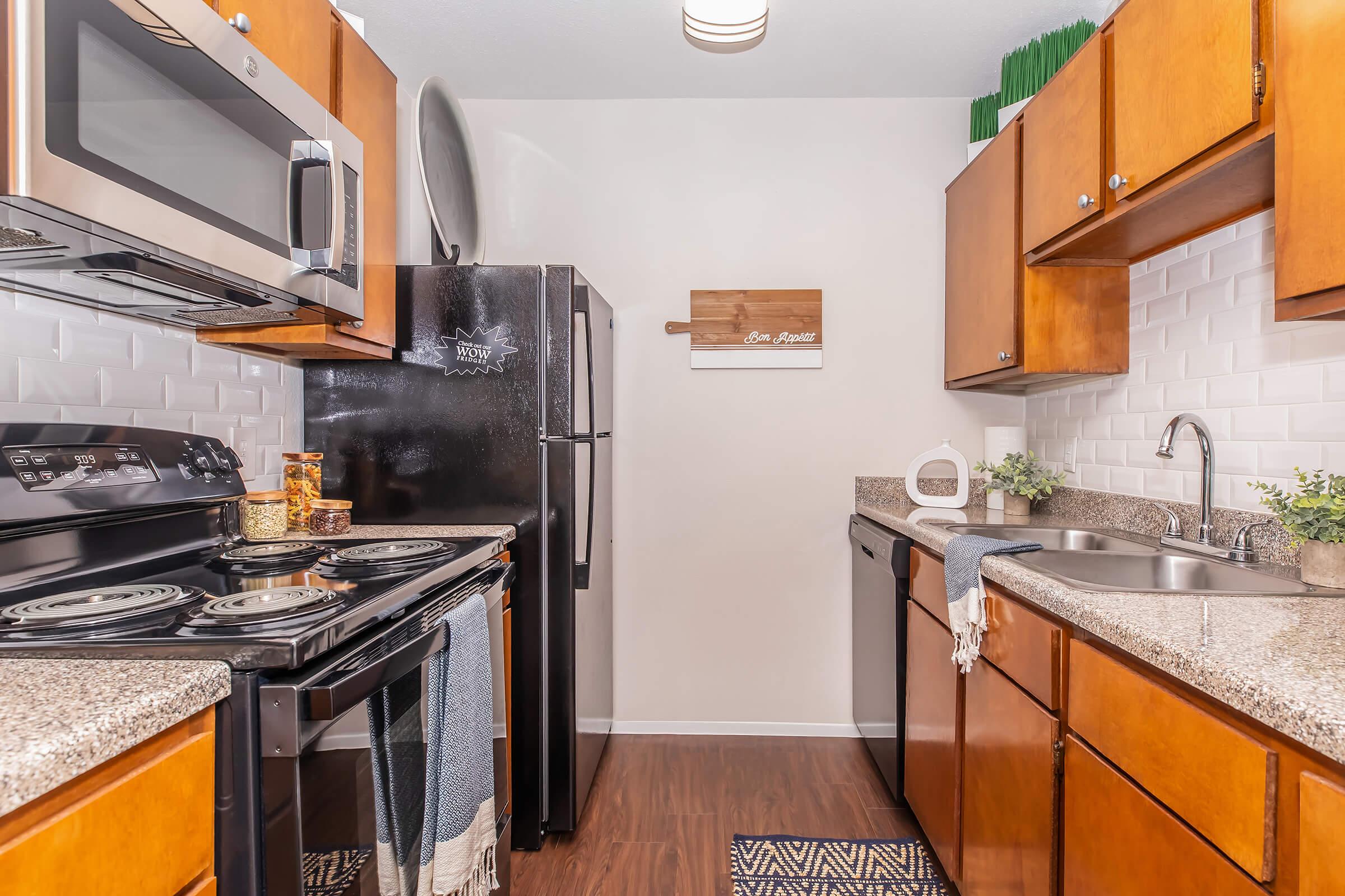 A modern kitchen featuring dark wooden cabinets, stainless steel appliances including a microwave and oven, and a black refrigerator. The counters are adorned with decorative items and a small plant. The backsplash is white subway tile, and there is a decorative wooden sign on the wall.