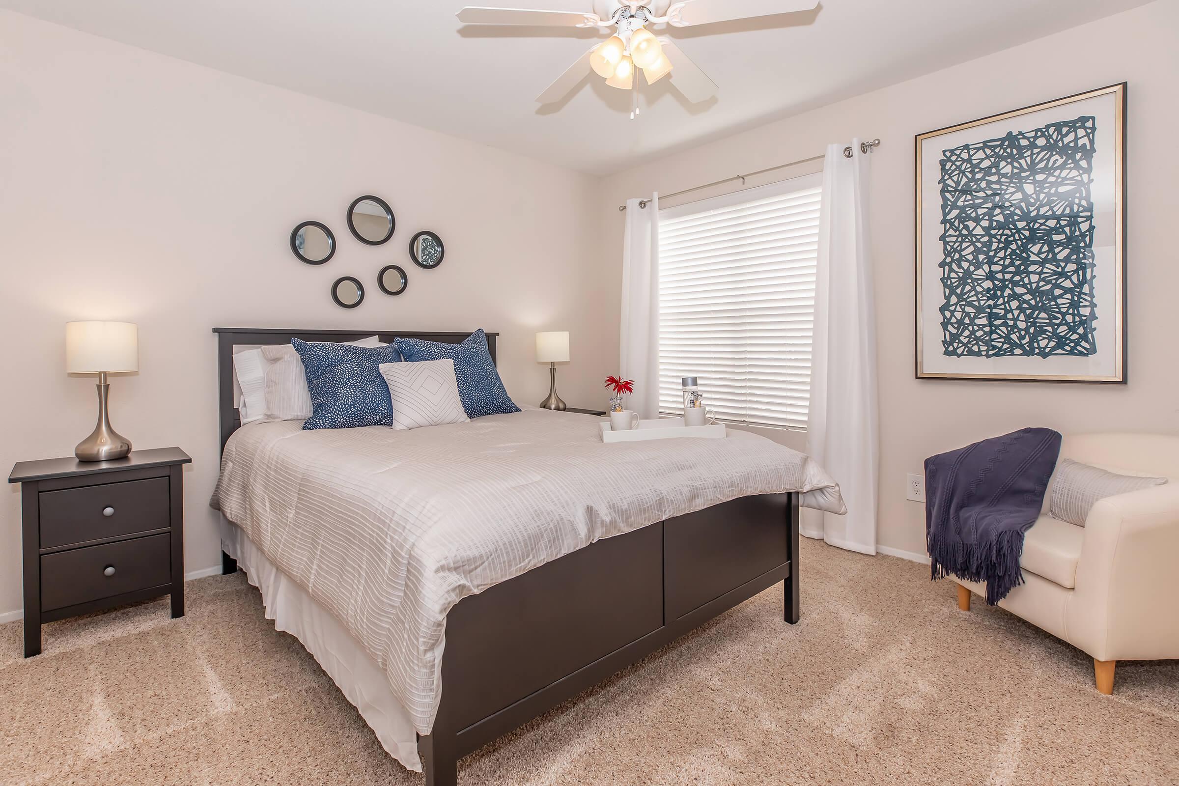 A cozy bedroom featuring a bed with gray and white bedding, a nightstand with lamps on either side, and decorative wall mirrors. A window with white curtains allows natural light to fill the room. A piece of abstract wall art adds a pop of color, and a small chair is positioned in the corner for relaxation.