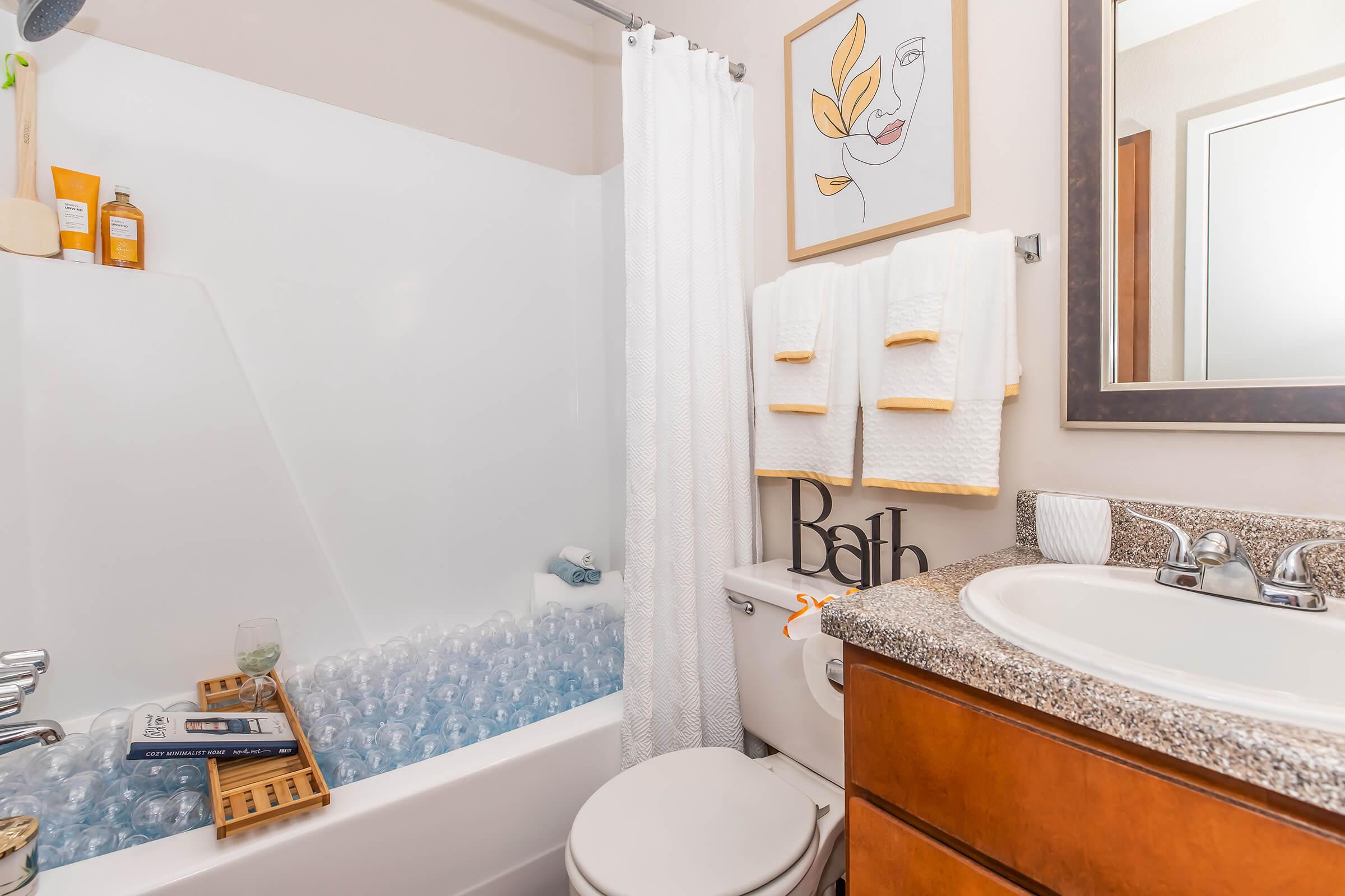 A stylish bathroom featuring a bathtub filled with blue bath balls, a shower curtain, beige walls, and wooden cabinetry. A white towel rack displays folded towels next to decorative items. A small glass sits beside a wooden bath tray with an open book, and a wall art piece adds a modern touch.