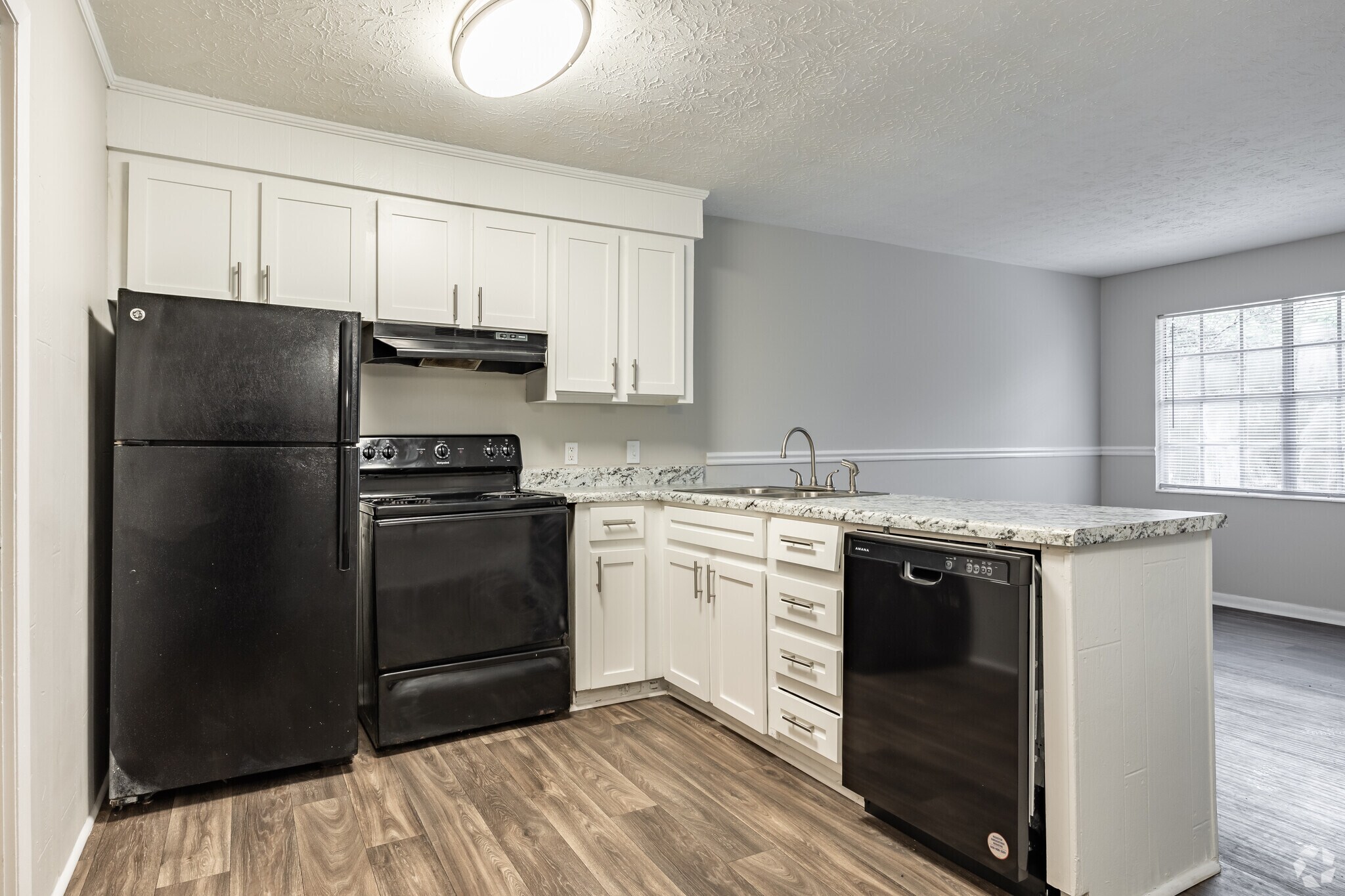 Modern kitchen with black appliances including a refrigerator, stove, and dishwasher. The cabinets are white with a granite countertop. The floor is wooden, and the walls are a light gray. A large window allows natural light to fill the space.