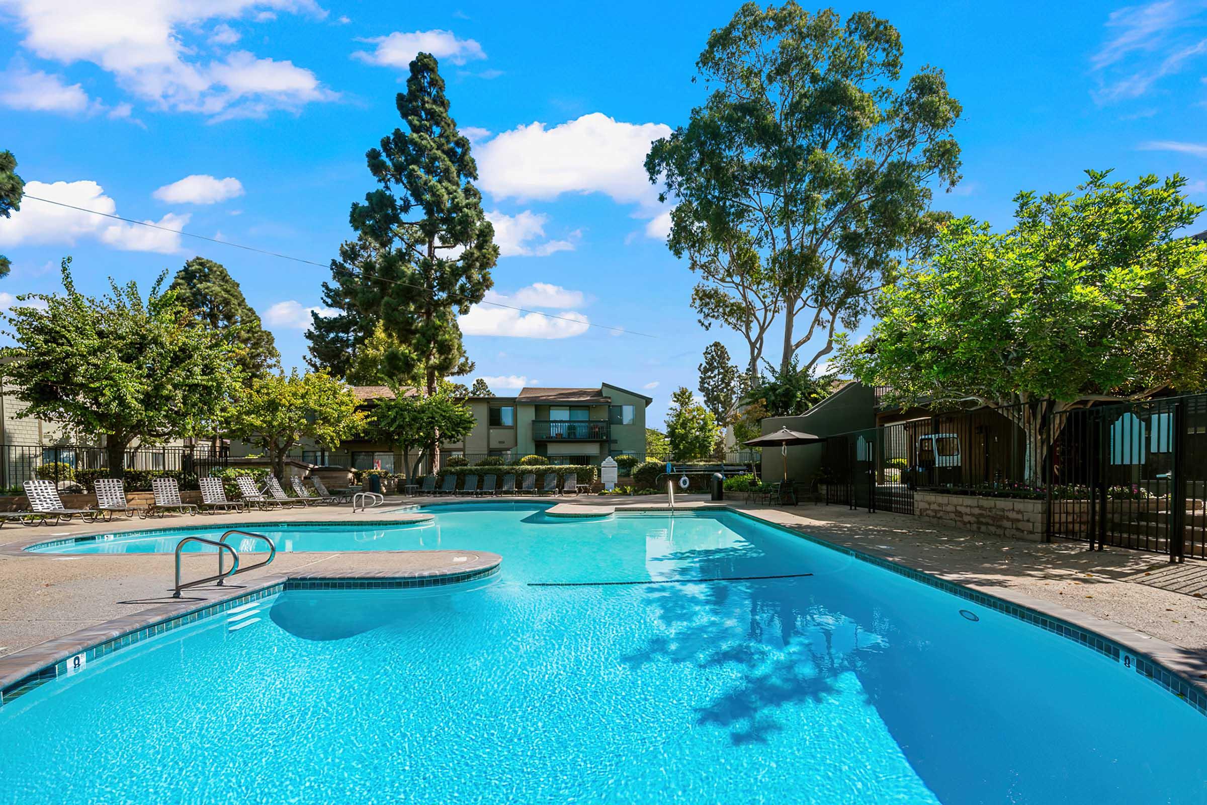 A clear blue swimming pool surrounded by lounge chairs, trees, and greenery. The sky is bright with fluffy white clouds, and there's a residential building in the background. The pool area is well-maintained, creating a relaxing outdoor atmosphere.