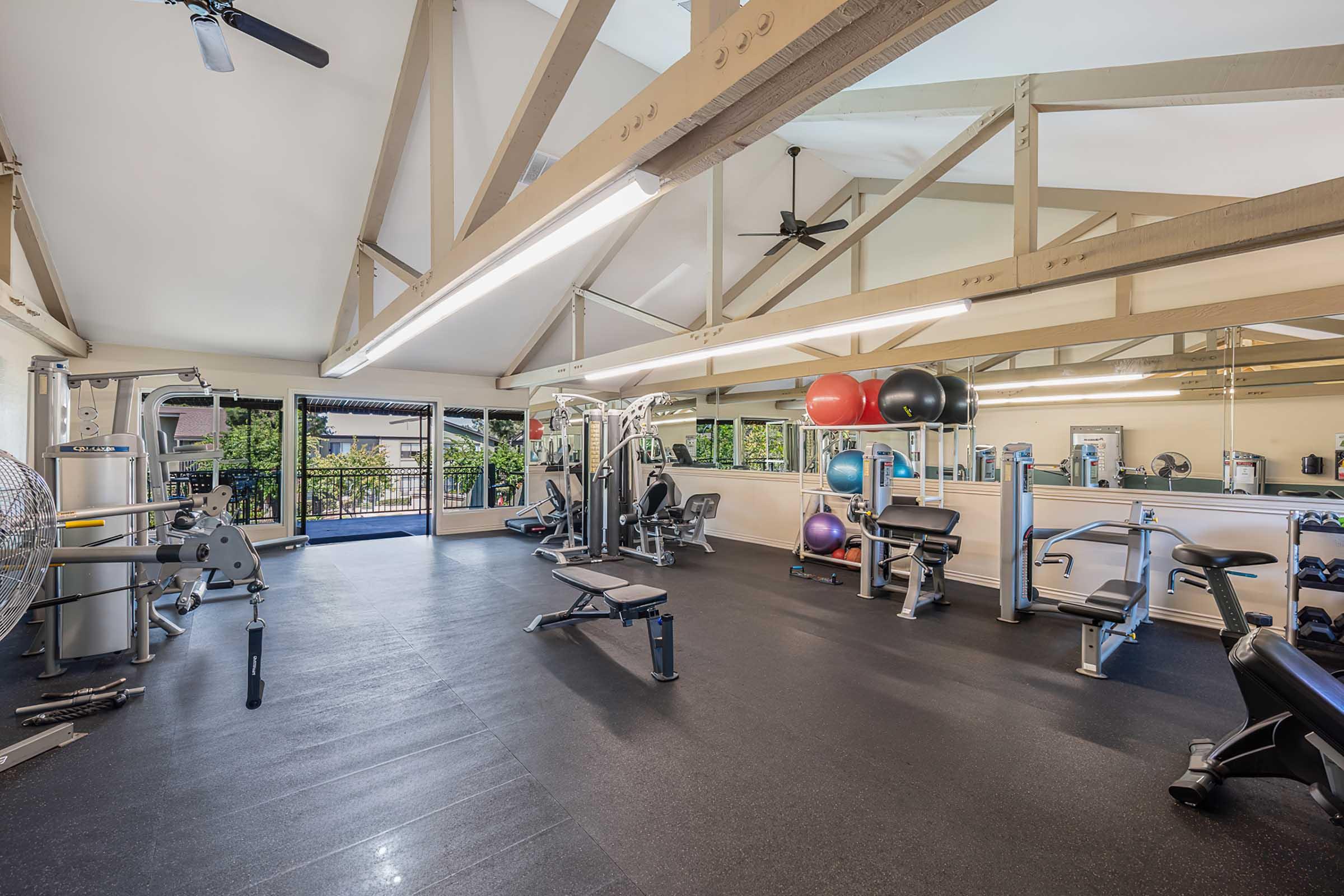 A well-lit gym featuring various exercise equipment including weight machines, benches, stability balls, and free weights. Large windows provide natural light and a view of greenery outside. The room has a high ceiling with exposed beams and ceiling fans for ventilation.