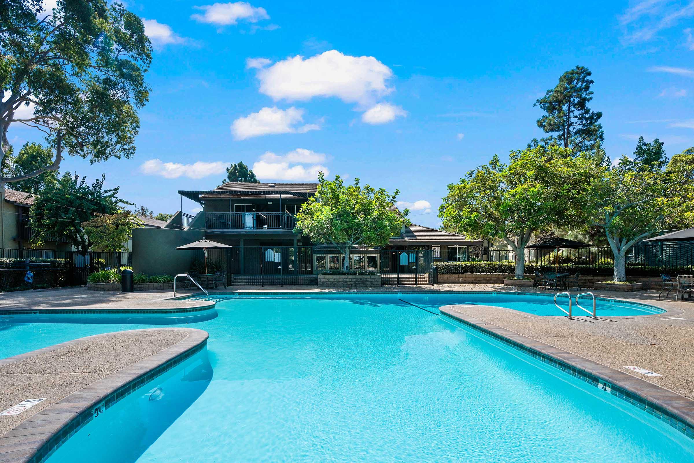 A clear blue swimming pool surrounded by lush green trees and well-maintained landscaping. In the background, there is a building with outdoor seating and umbrellas, under a bright blue sky with a few fluffy clouds. The pool area invites relaxation and leisure.