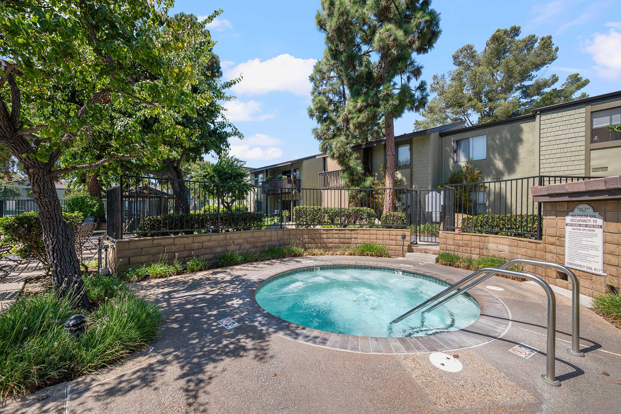 A sparkling hot tub surrounded by well-maintained greenery and trees, with an apartment building visible in the background. The area is enclosed with a fence, and the sky is clear with fluffy clouds.