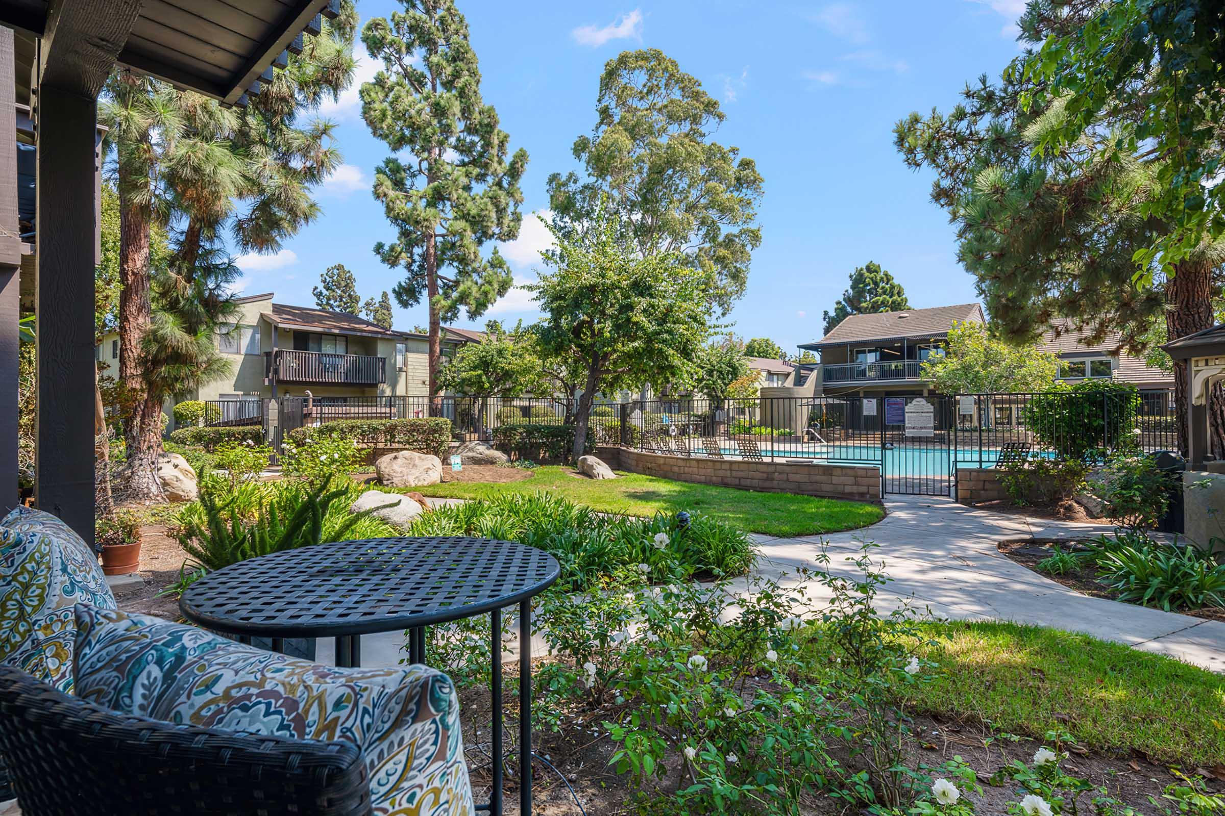 A serene outdoor view from a patio featuring a small round table and chairs surrounded by lush greenery. In the background, a sparkling pool is visible, framed by buildings and trees, creating a tranquil, inviting atmosphere.